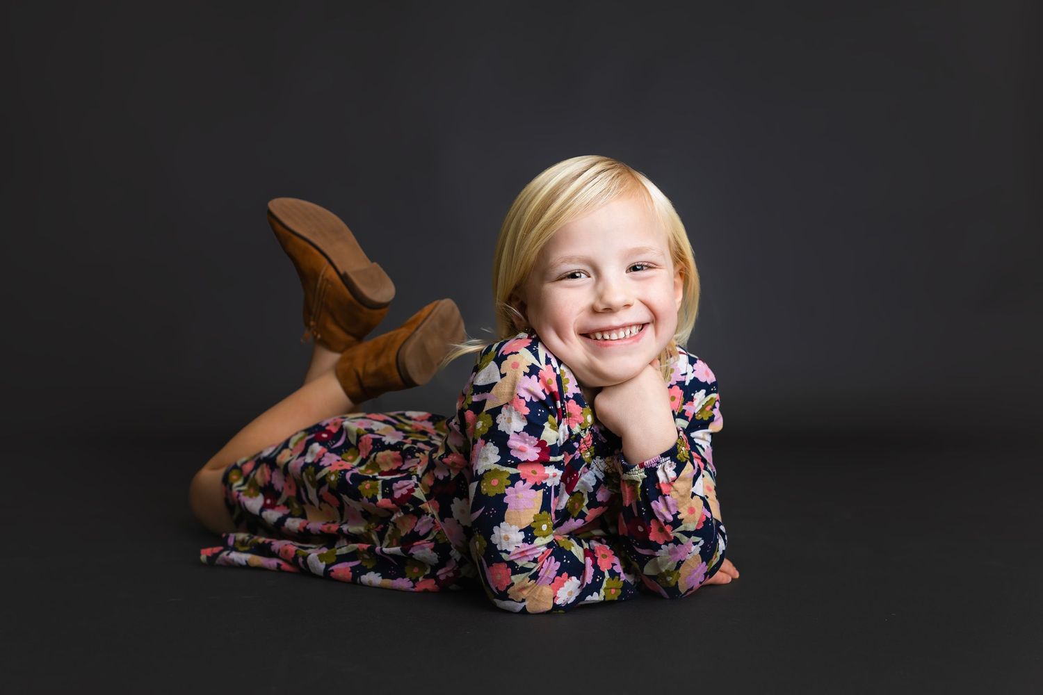 Adorable blonde girl in floral dress lying on floor smiling with chin resting on hands in studio portrait.