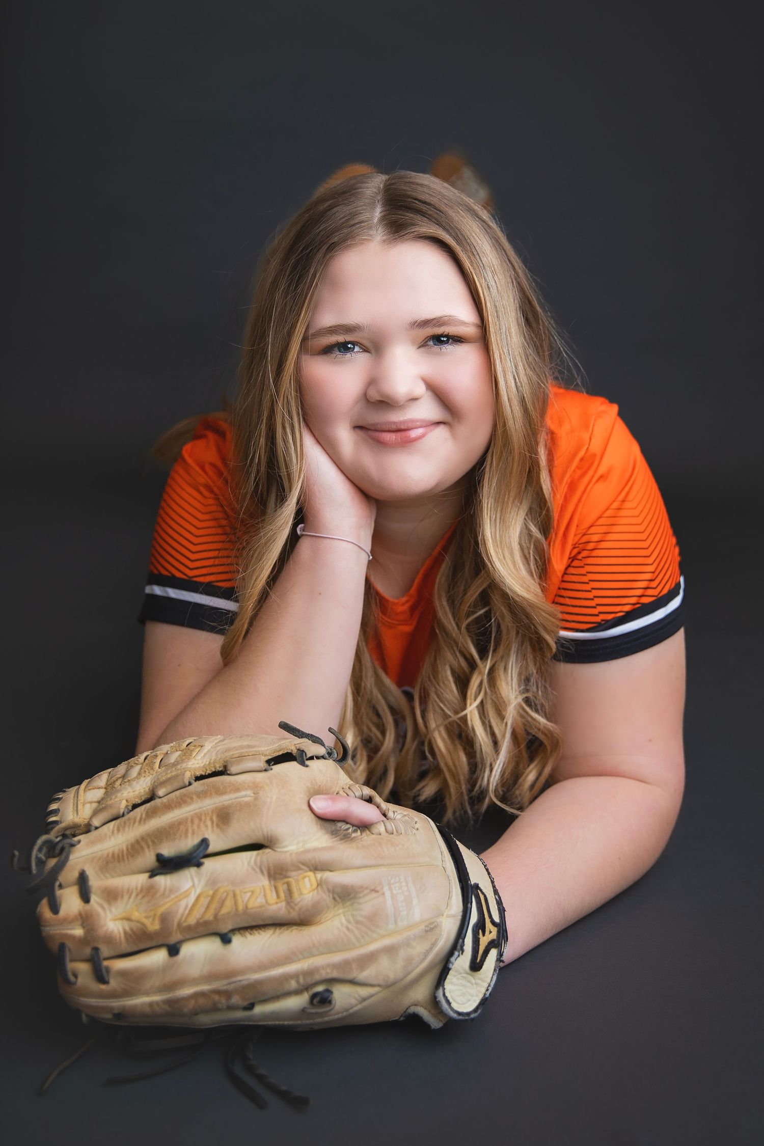 Smiling young softball player in orange jersey poses with glove in relaxed studio portrait.