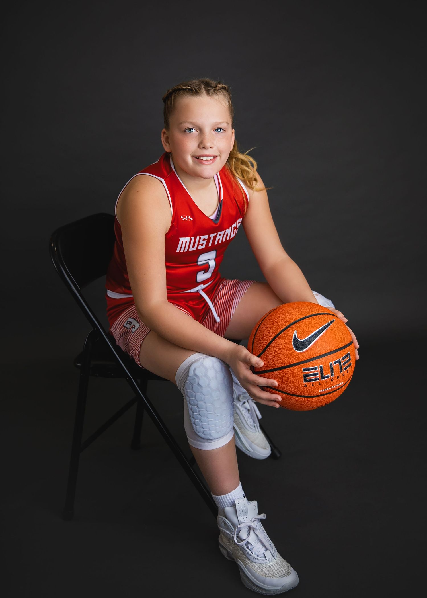 Young female basketball player in red Mustangs uniform sitting on chair holding Nike Elite basketball.