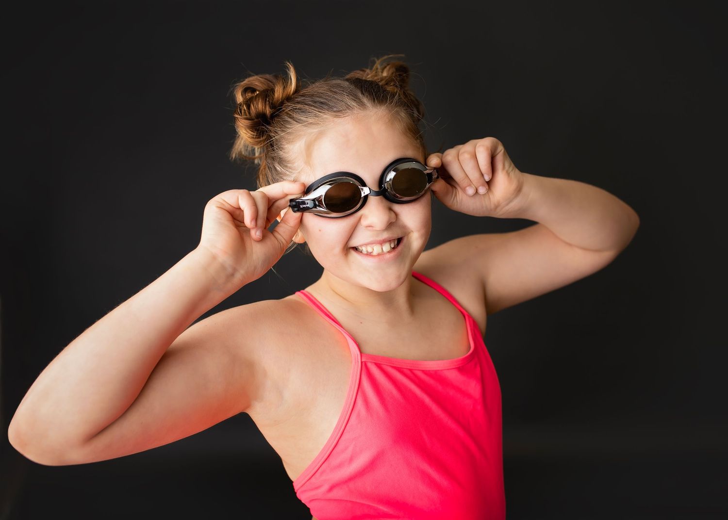 Smiling young girl in pink swimsuit adjusting swim goggles against a dark background.