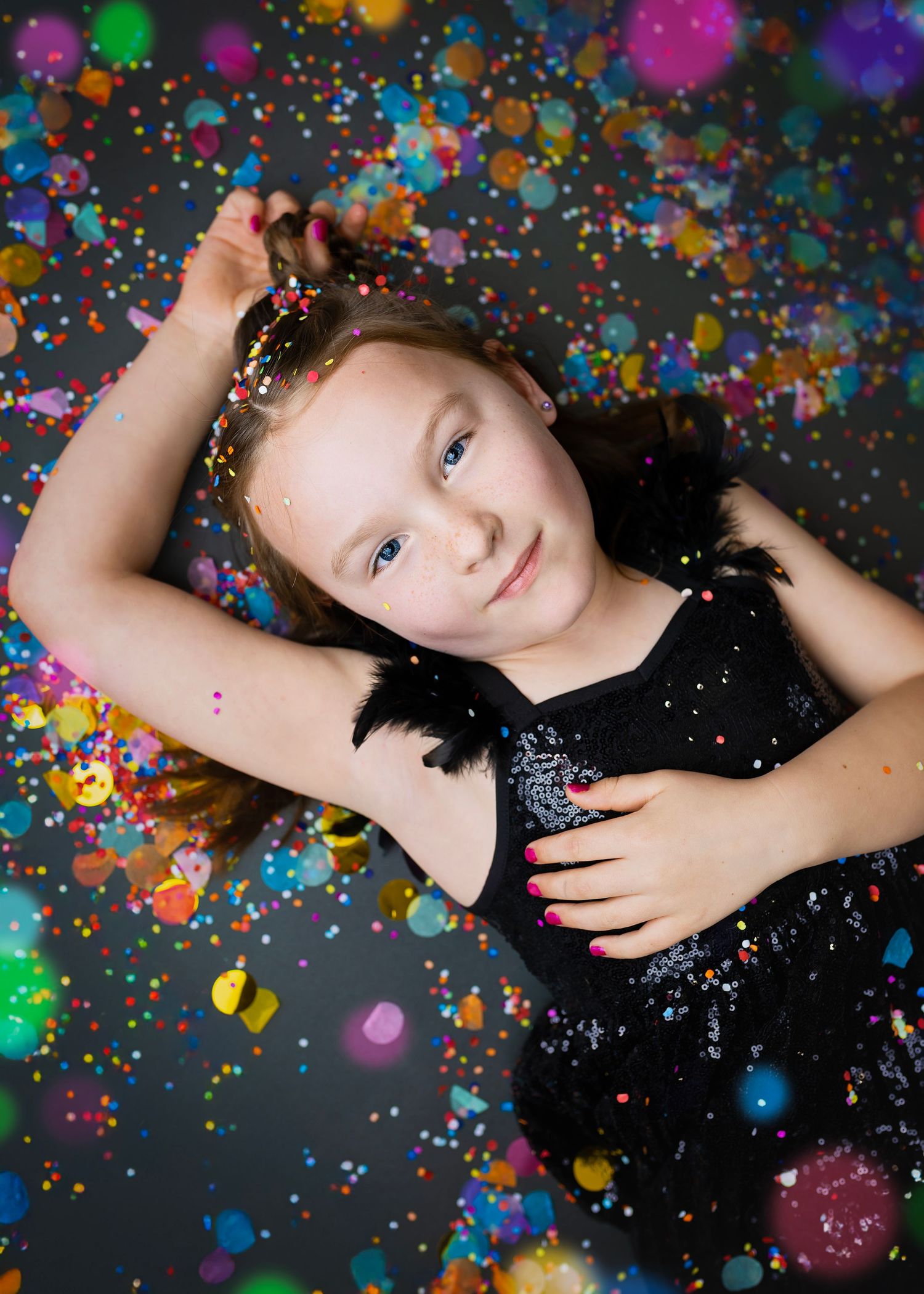 Smiling girl in black sequin dress lying amid colorful confetti with pink nails.