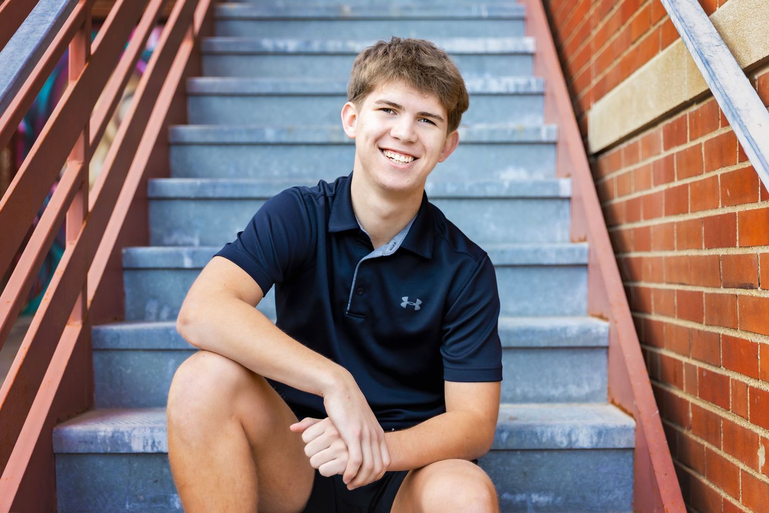 Smiling teenage boy in navy polo shirt sitting on brick steps outdoors.