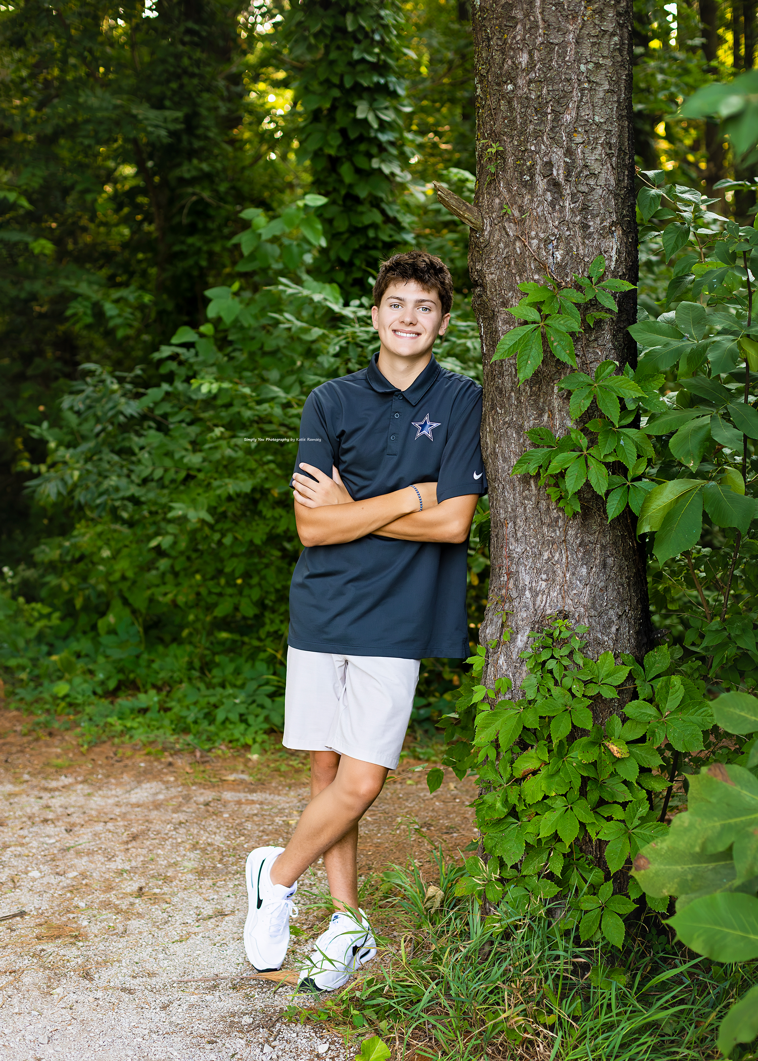 Smiling teen boy in navy polo and gray shorts leans against tree on wooded path with arms crossed.