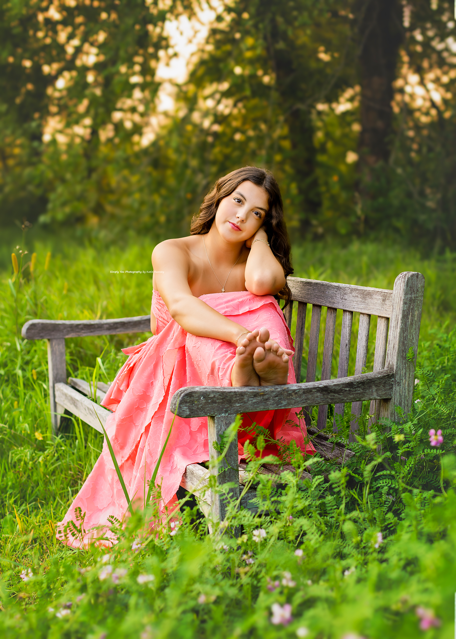 Young woman in pink gown seated on rustic wooden bench surrounded by lush green meadow with wildflowers at golden hour.