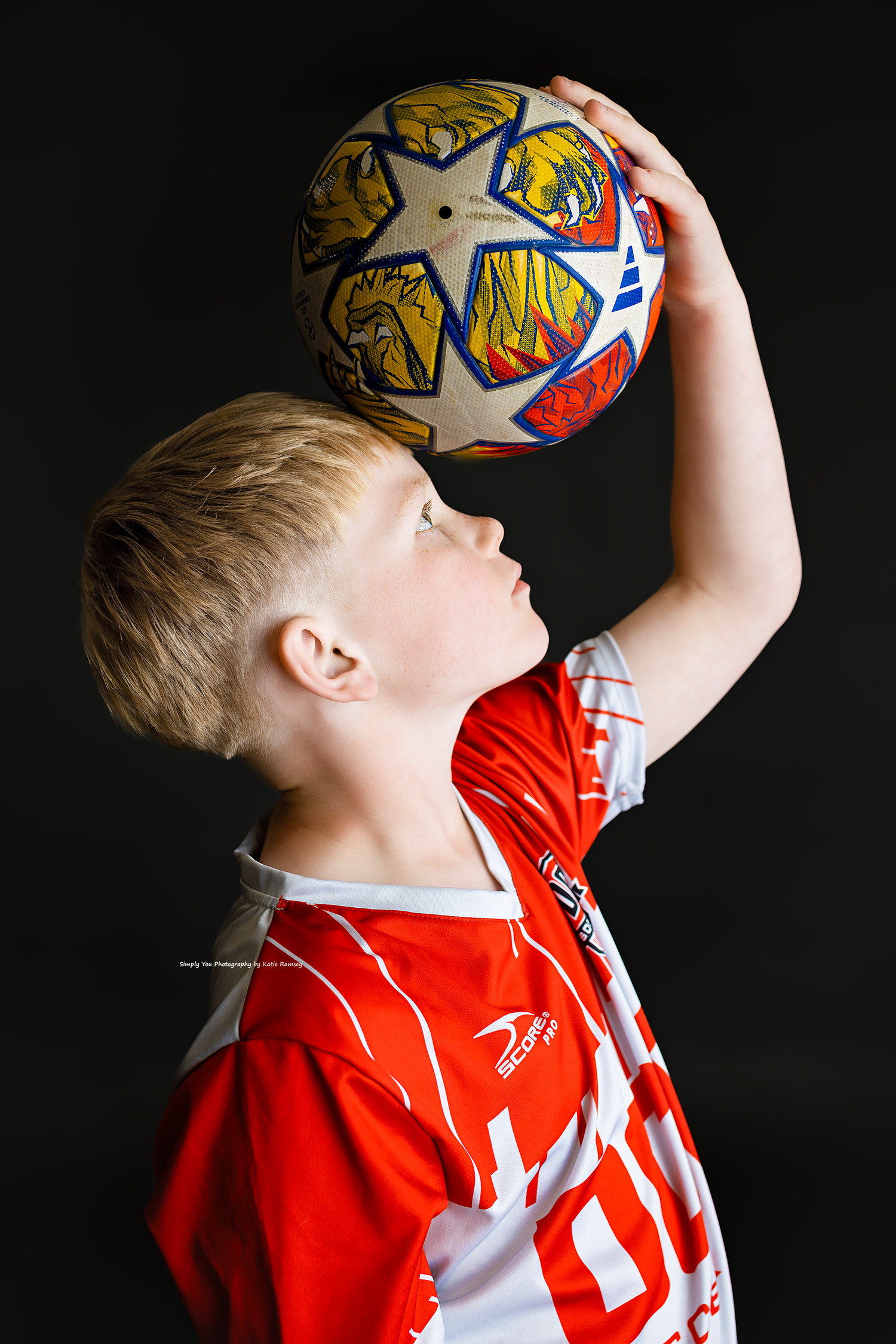 Young blonde boy in red soccer jersey holds a colorful Champions League ball above his head against black background.