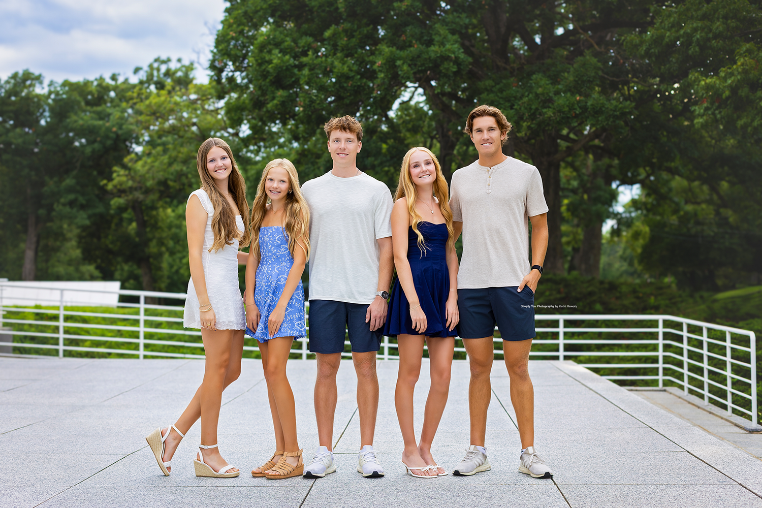 Five smiling teenagers posing together outdoors on a sunny day, dressed in casual summer outfits.