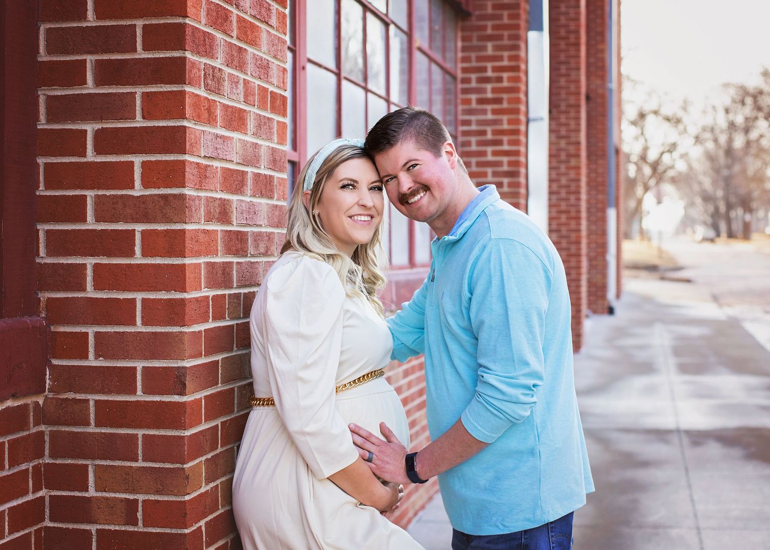 Smiling pregnant couple leans against a brick wall, with man embracing woman in white maternity dress.