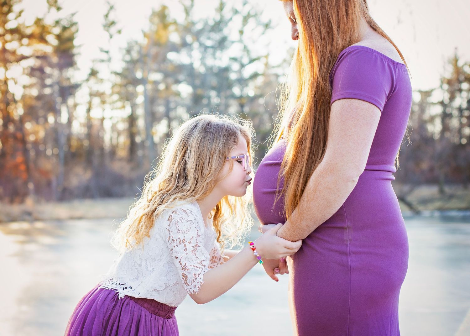 Young girl in white lace top kisses pregnant mothers purple-dress-clad baby bump near a sunlit lake.