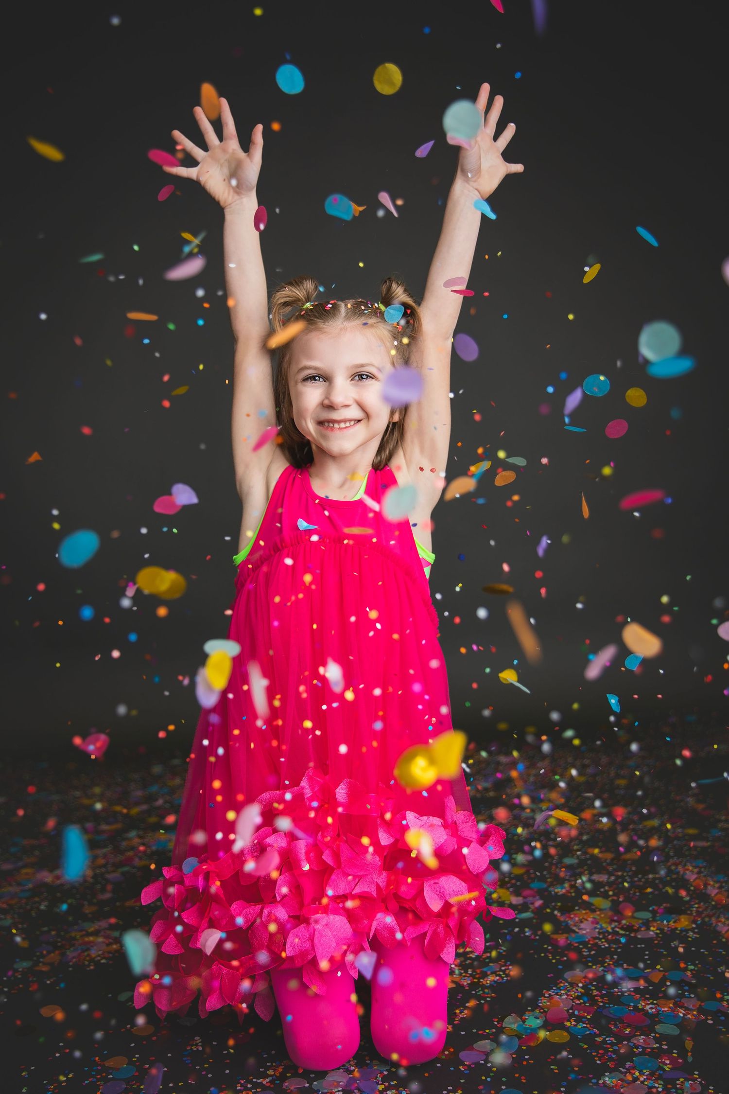 Happy girl in red dress with arms raised surrounded by colorful confetti on dark background.