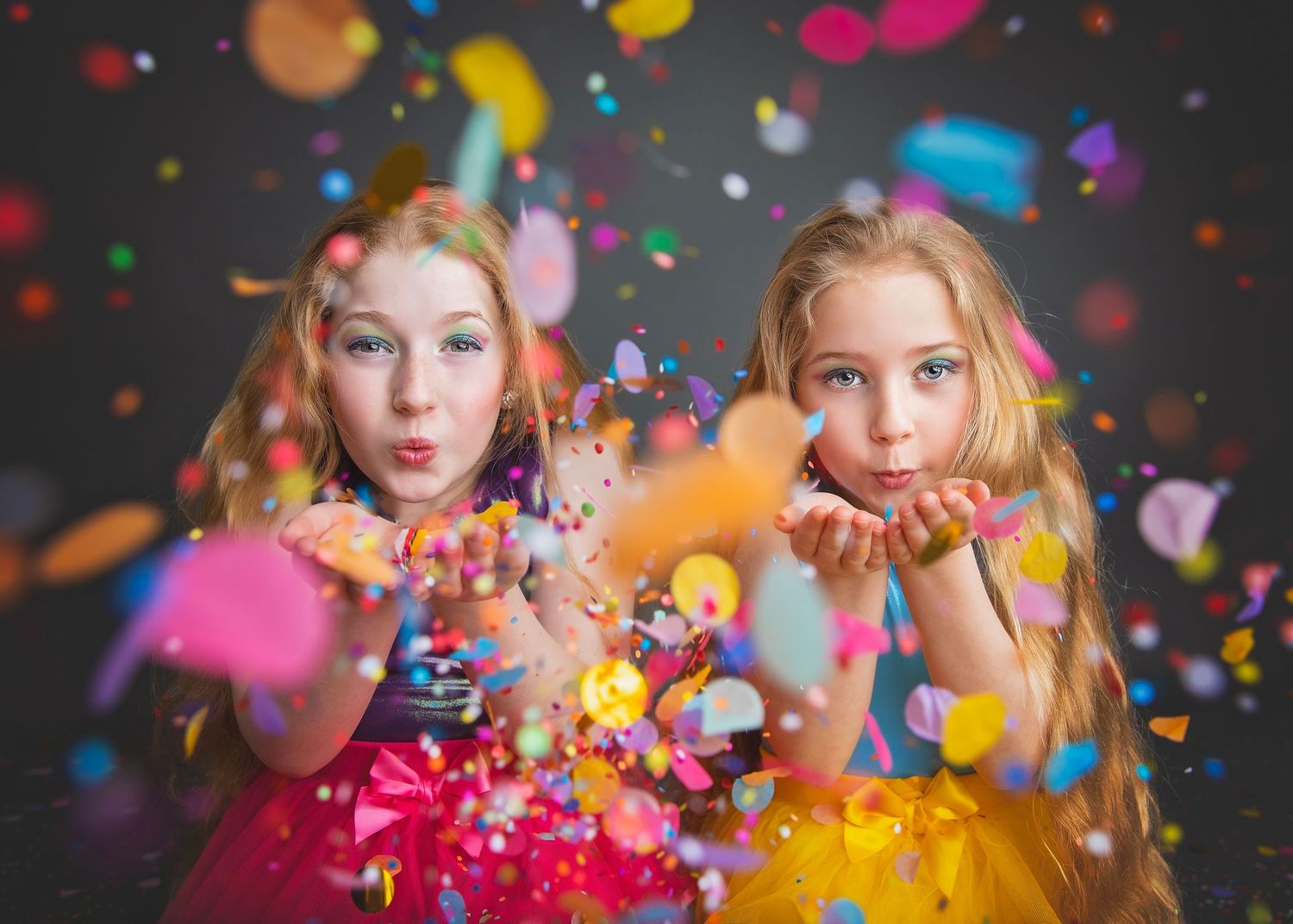 Two young girls blowing colorful confetti toward camera in a festive studio photo session.