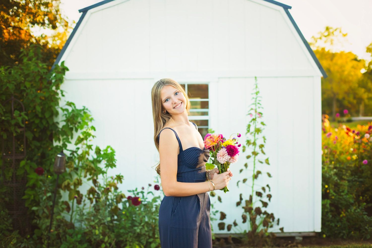 Smiling blonde woman in navy jumpsuit holds colorful flower bouquet near white barn outdoors.