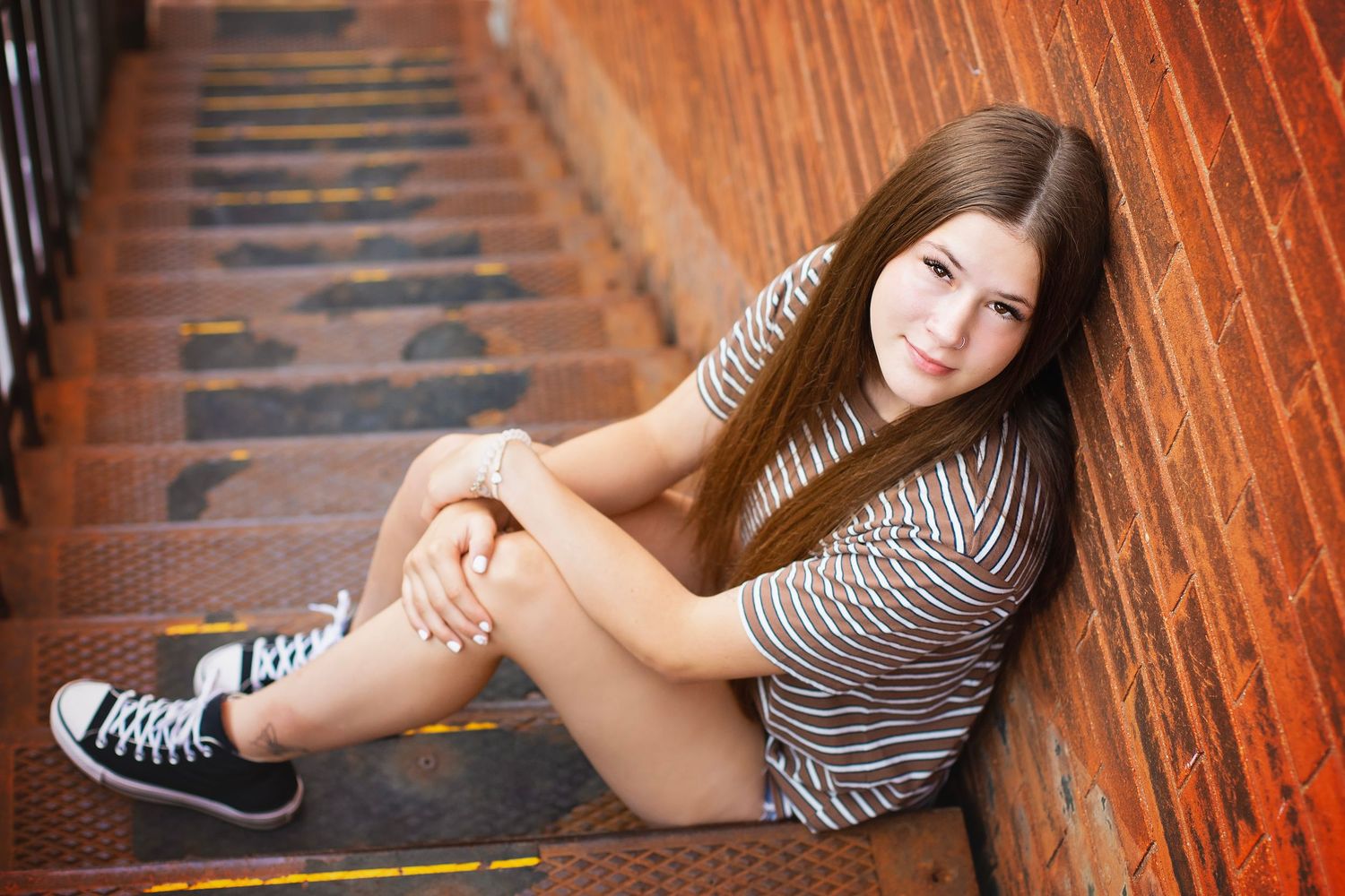 Teenage girl in striped shirt sitting on brick staircase wearing black sneakers smiling at camera.