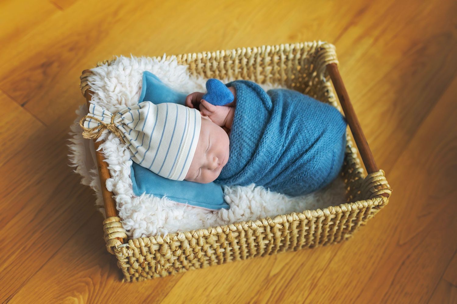 Sleeping newborn baby swaddled in blue wrap wearing striped hat resting in wicker basket on white fur.