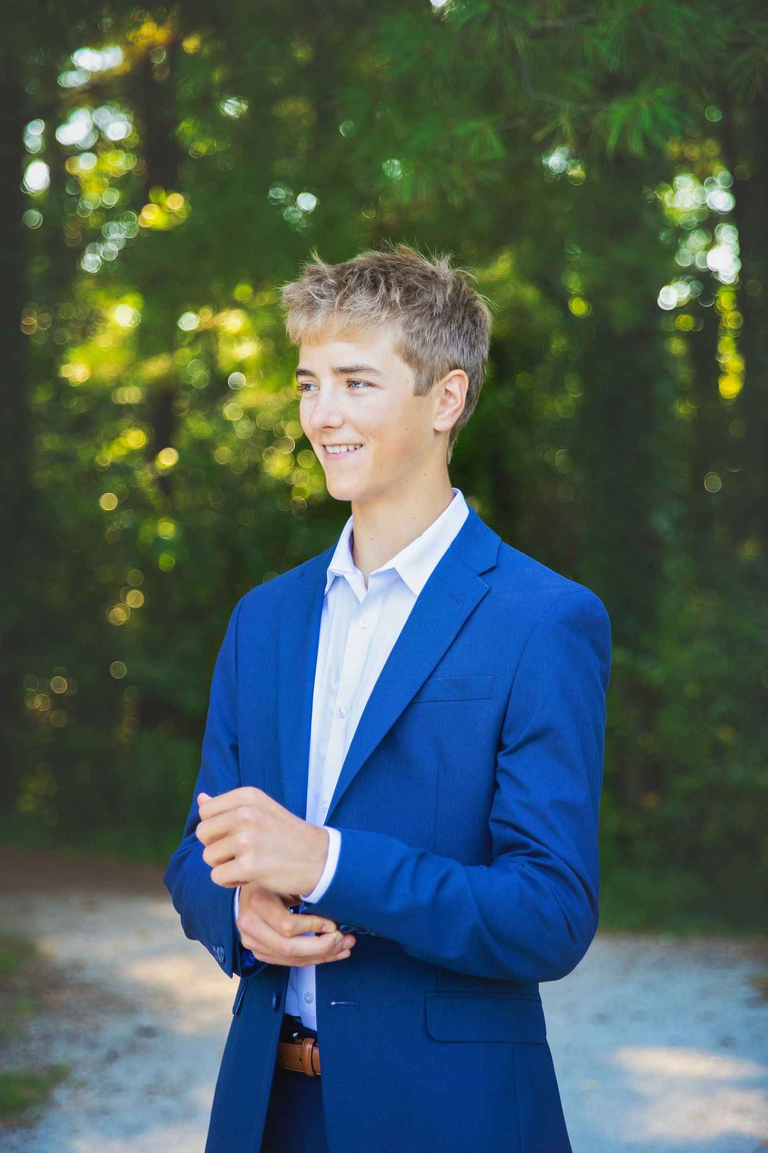 Smiling teenage boy in blue blazer and white shirt standing on forest path adjusting his sleeve.