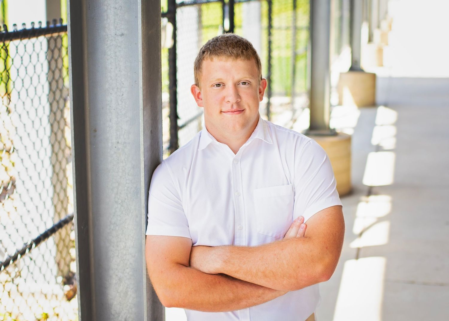 Young man in white polo shirt stands with arms crossed near a chain-link fence outdoors.