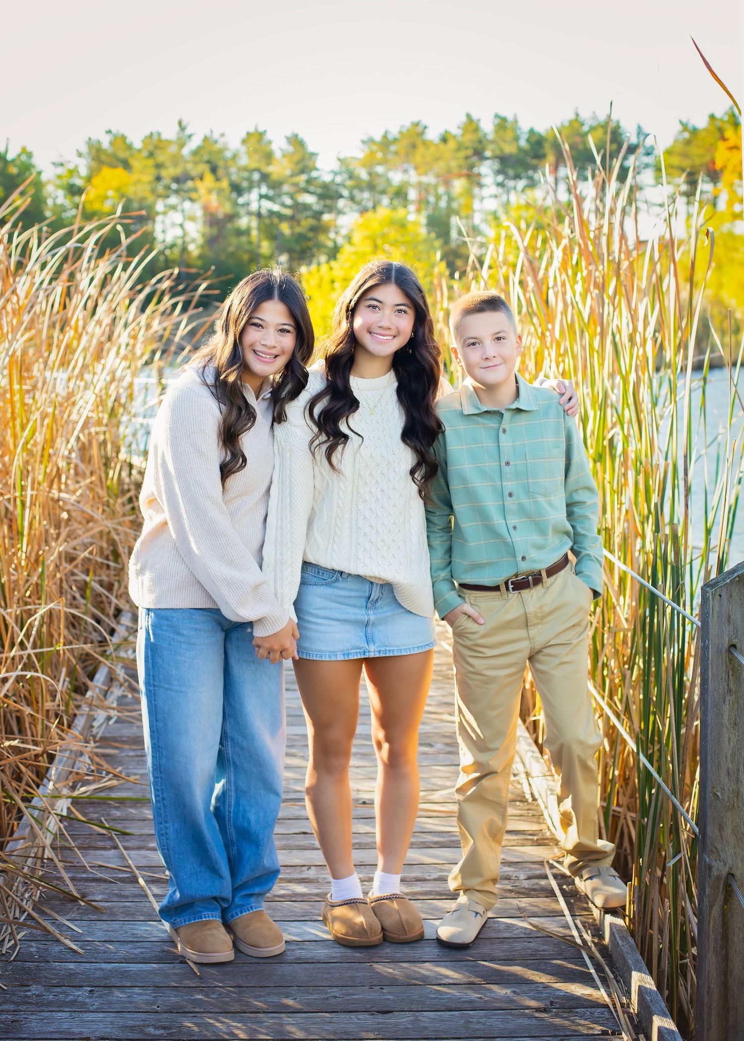 Three siblings posing on a wooden dock surrounded by tall reeds at sunset.