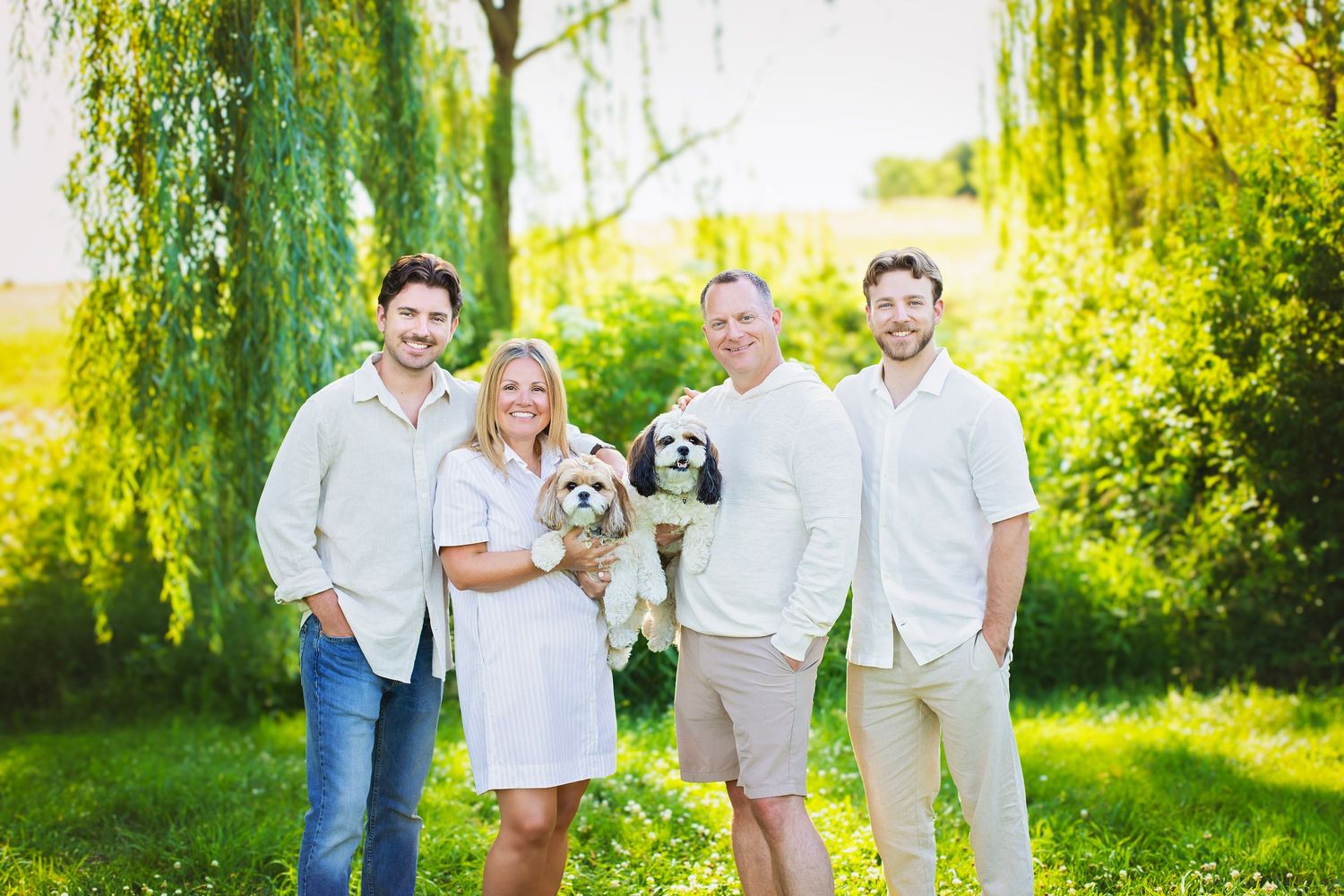 Smiling family of four poses outdoors under a weeping willow tree holding two small fluffy dogs.