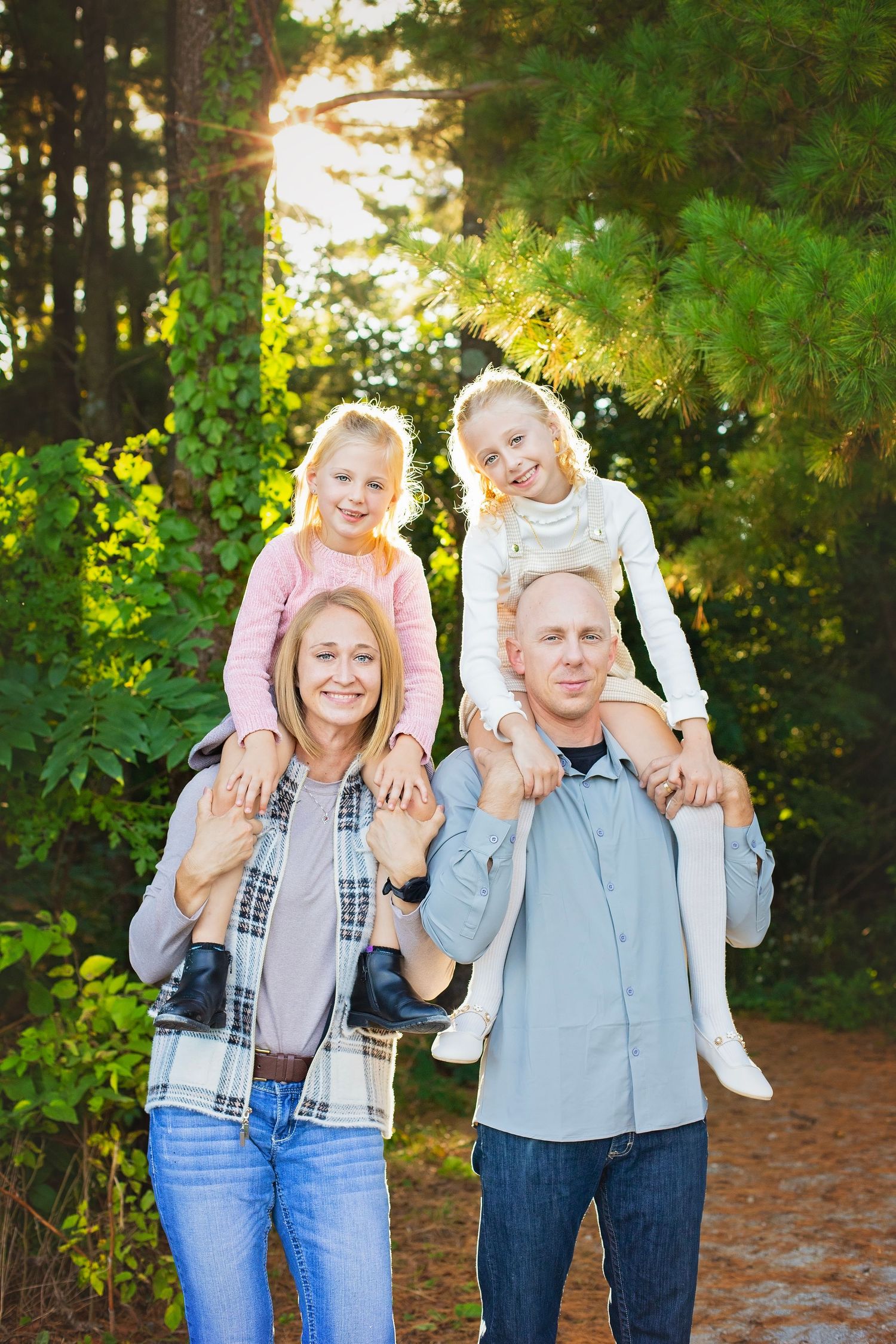 Parents carrying two young daughters on their shoulders in a sunny green forest setting.