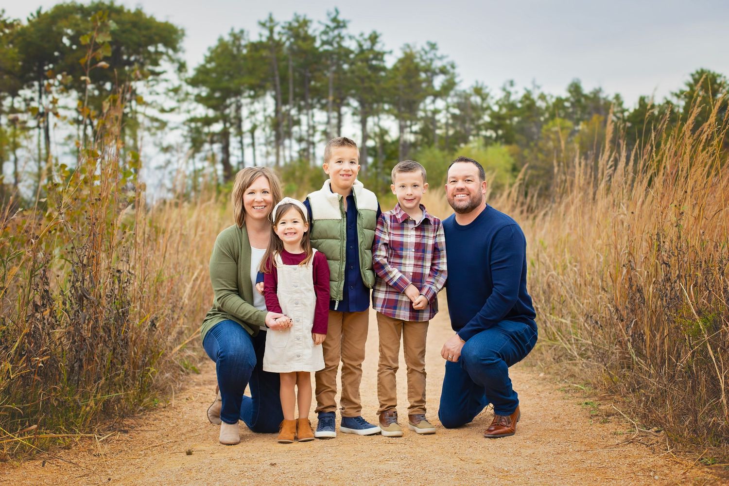 A family of five poses on a scenic dirt path surrounded by tall grasses and trees in autumn.