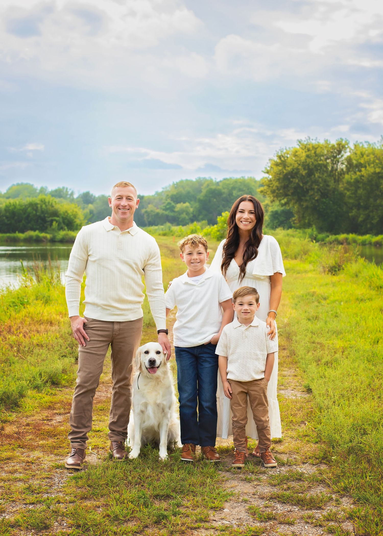 Family of four with golden retriever posing on a scenic green path near a lake on a cloudy day.