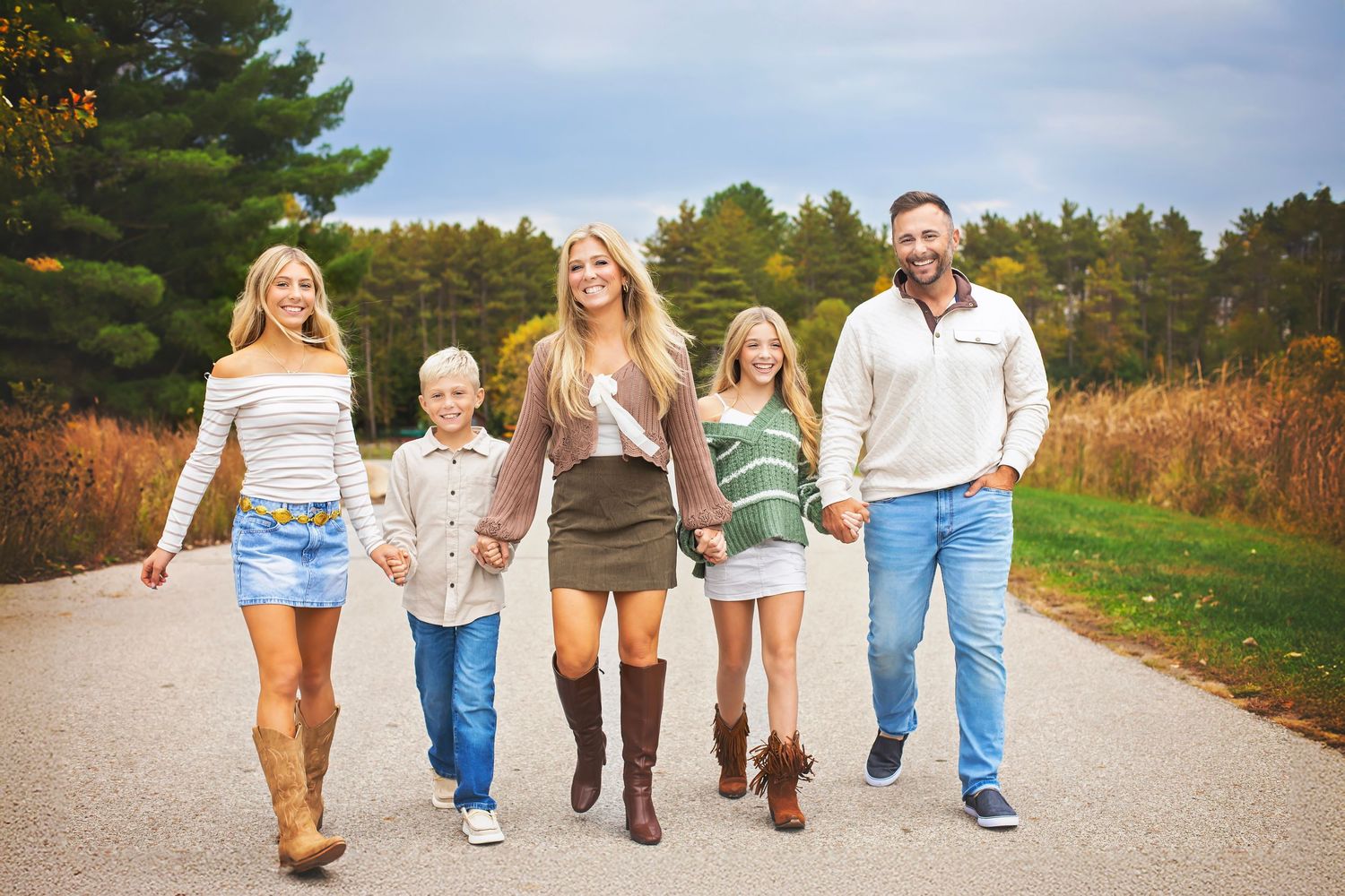 A happy family of five walks hand-in-hand down a scenic autumn road lined with colorful fall foliage.