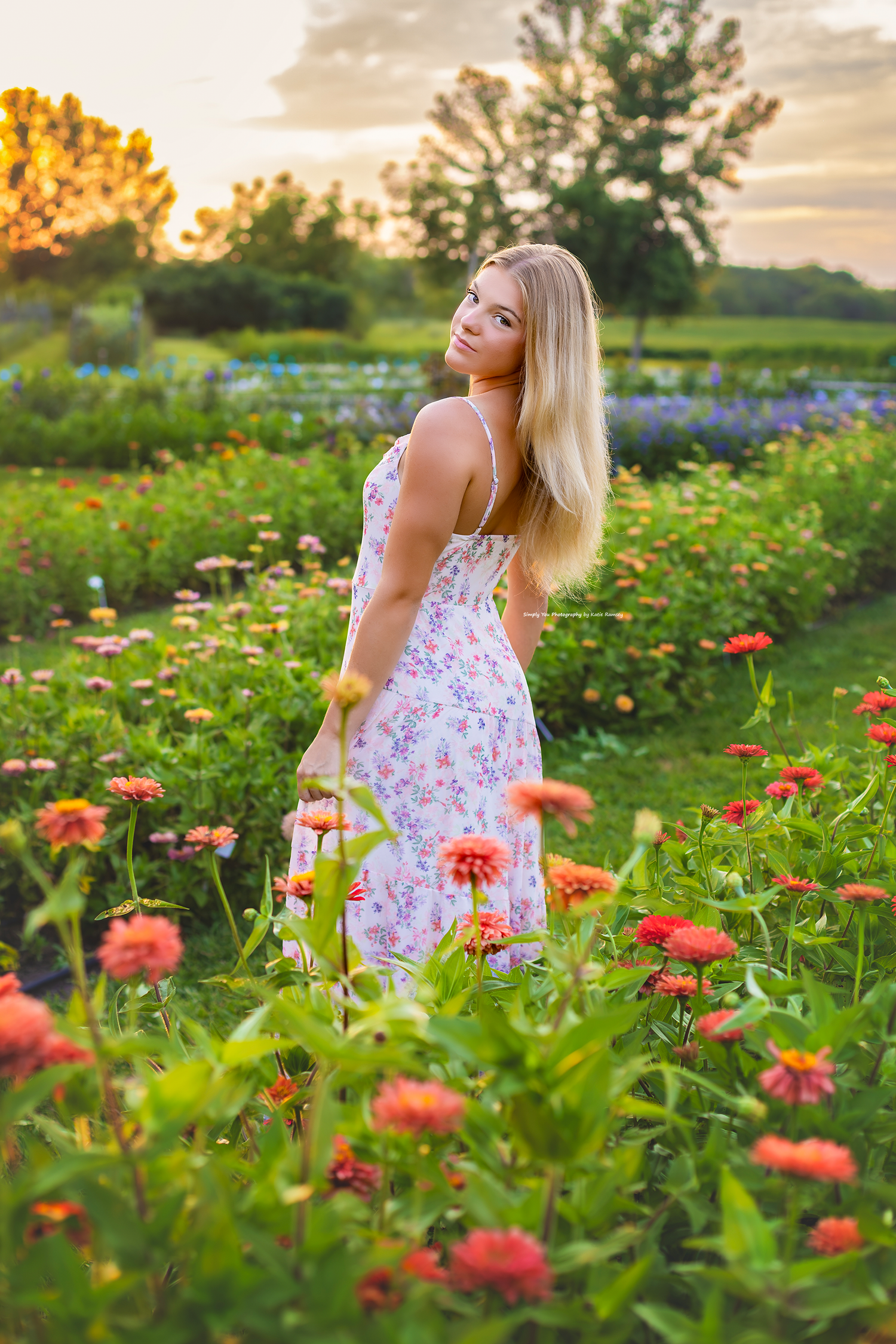 Blonde woman in floral sundress standing among colorful zinnia flowers in a garden at golden hour sunset.