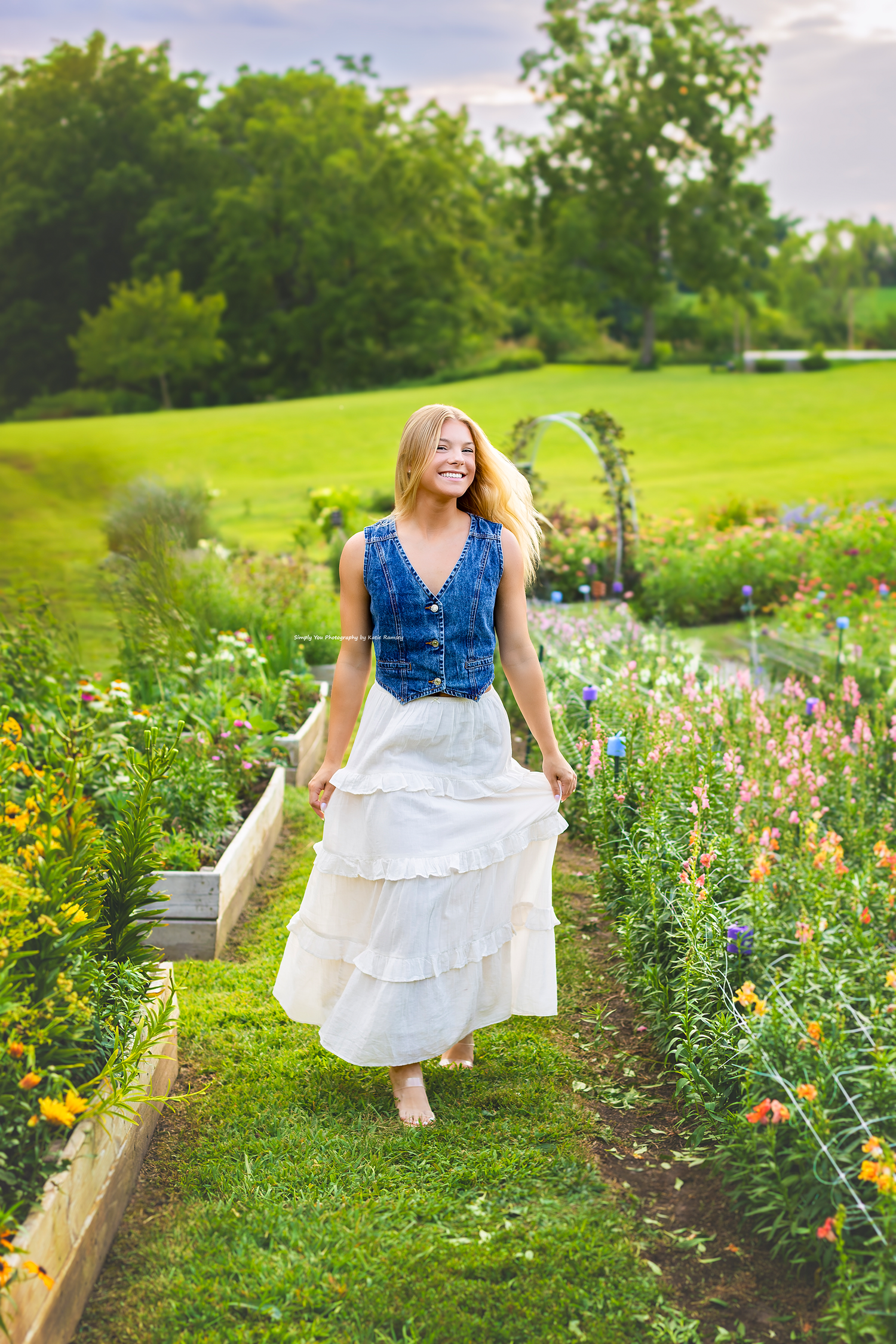 Young woman in denim vest and white ruffled skirt walking through a lush garden path with colorful flowers.