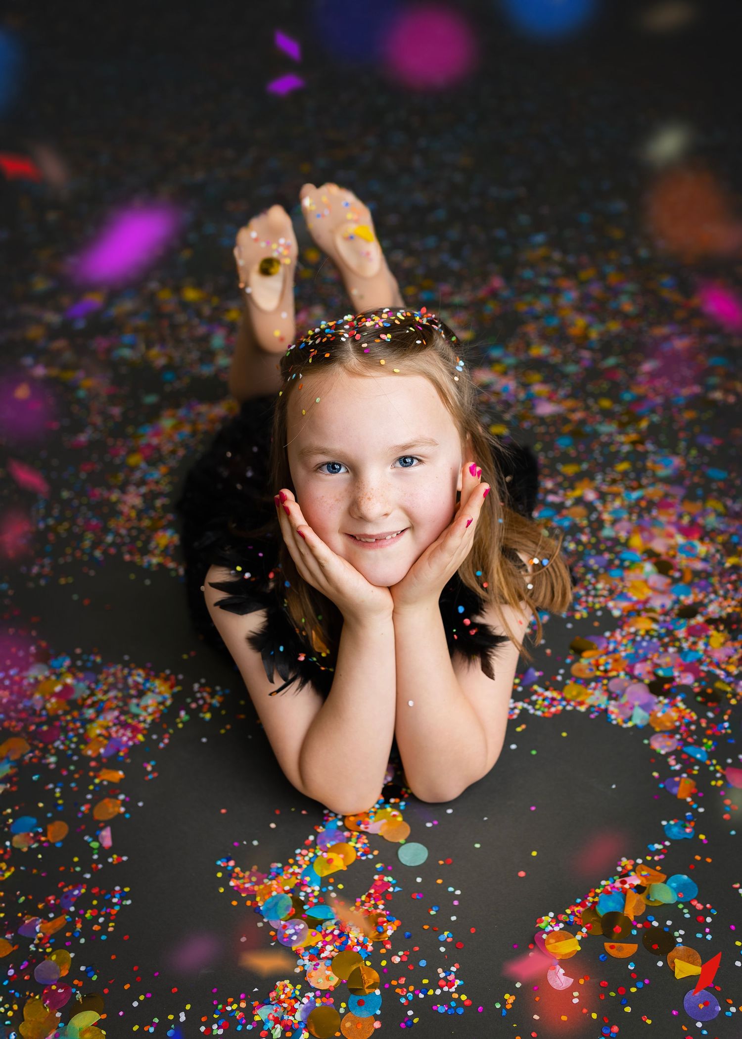 Cheerful girl in black dress lying on floor surrounded by bright colorful confetti.