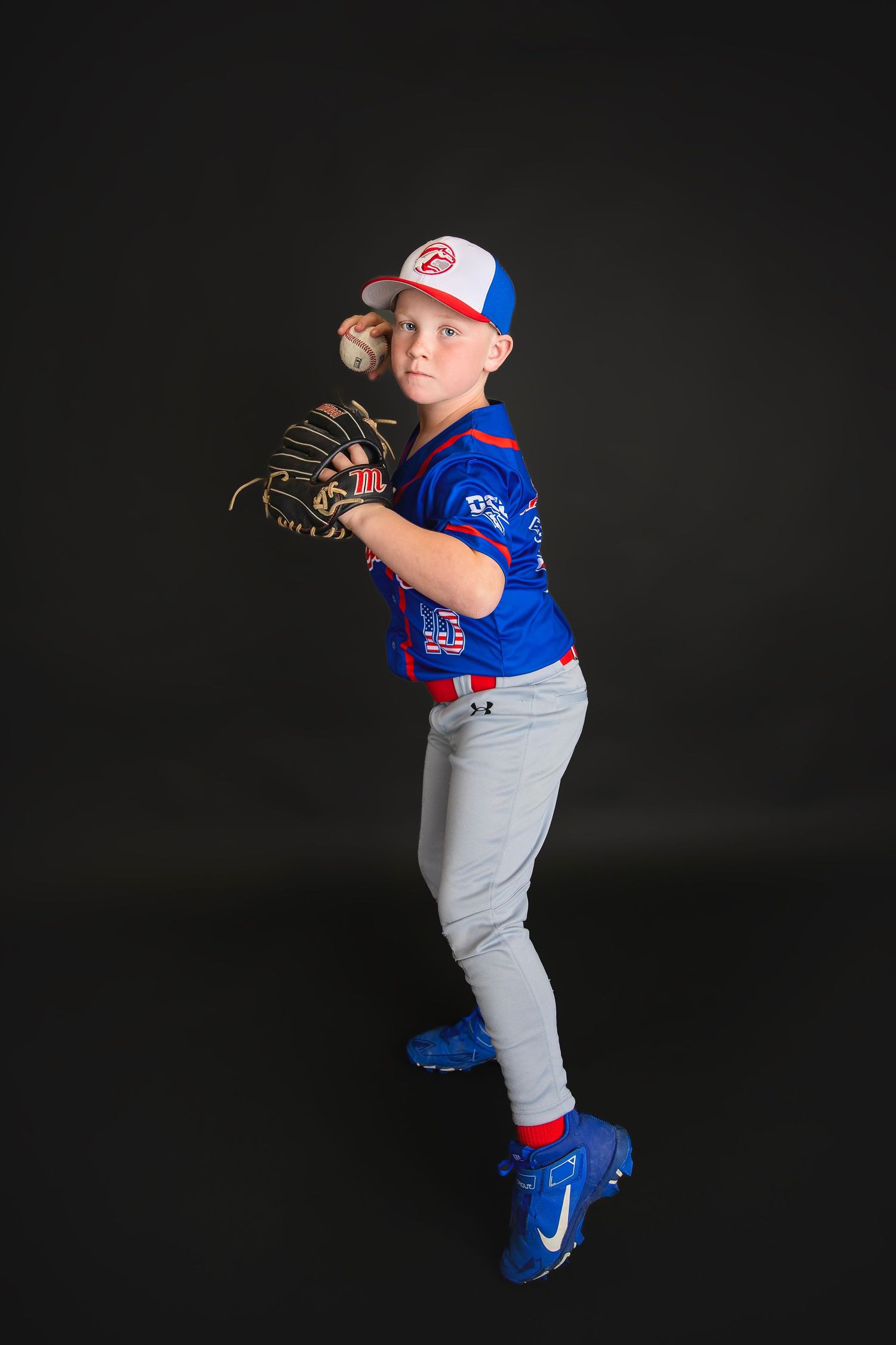 Young boy in blue Mustangs baseball uniform posing in pitching stance with glove and ball.