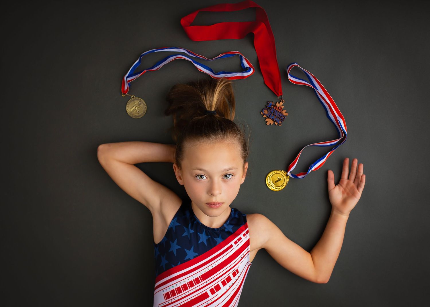 Young gymnast lying down surrounded by gold medals and colorful ribbons on dark background.