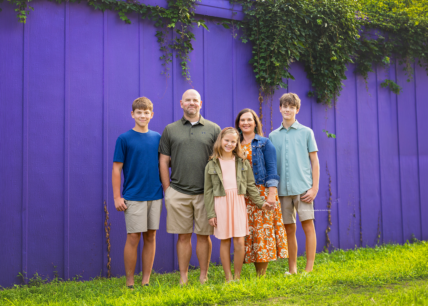 Family of five posing and smiling in front of a vibrant purple wooden fence with green ivy.