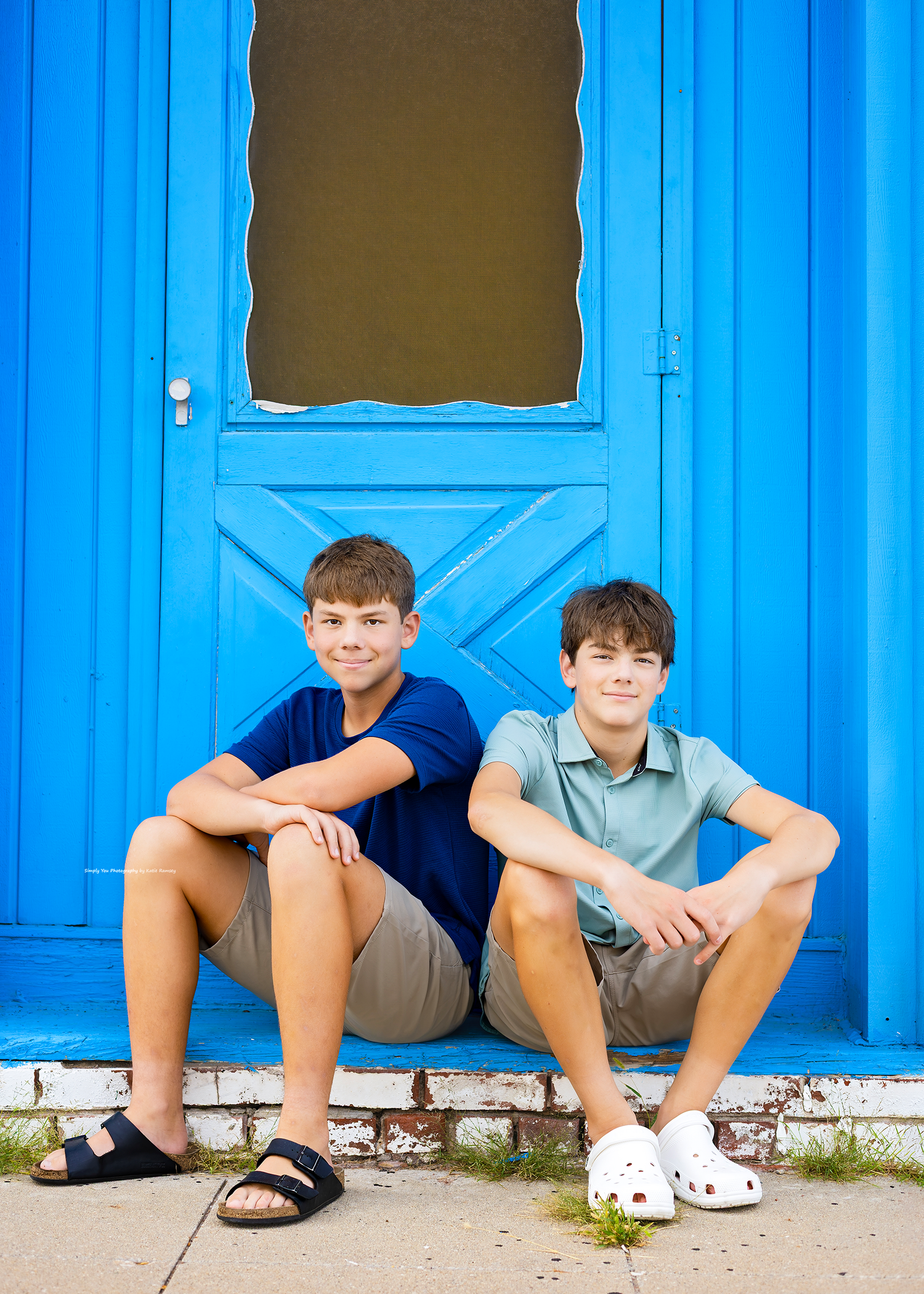Two smiling boys sitting in front of a bright blue door, wearing casual summer outfits and sandals.