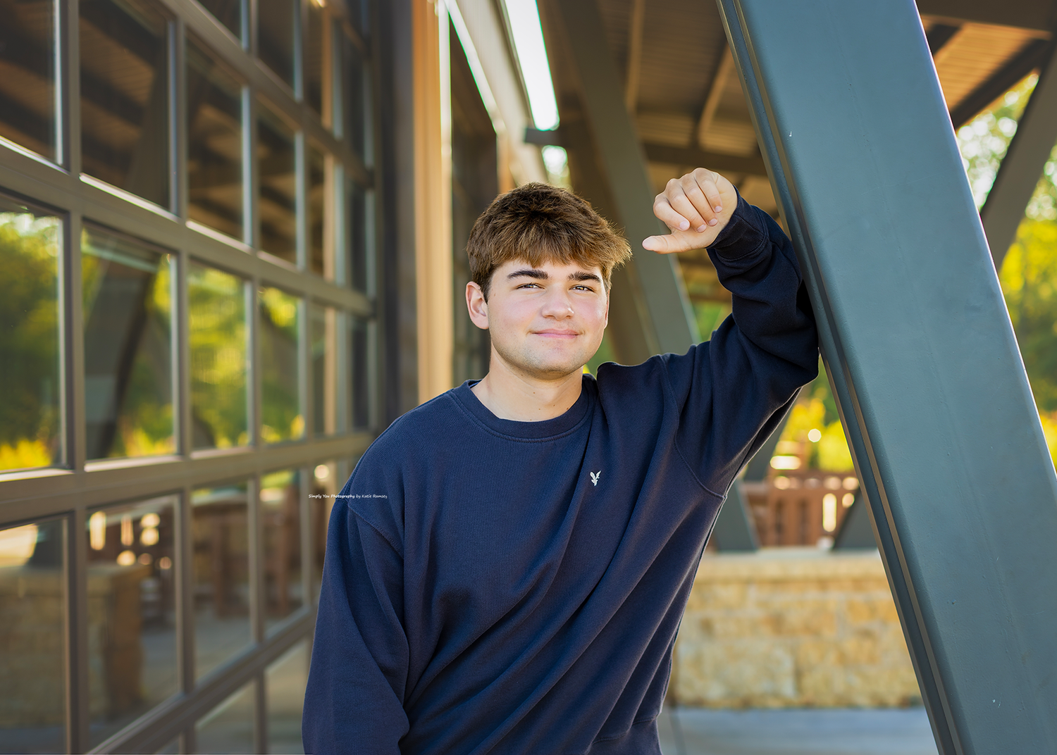Teen boy in navy sweatshirt leaning against a metal beam outdoors, smiling confidently in a senior portrait photo.