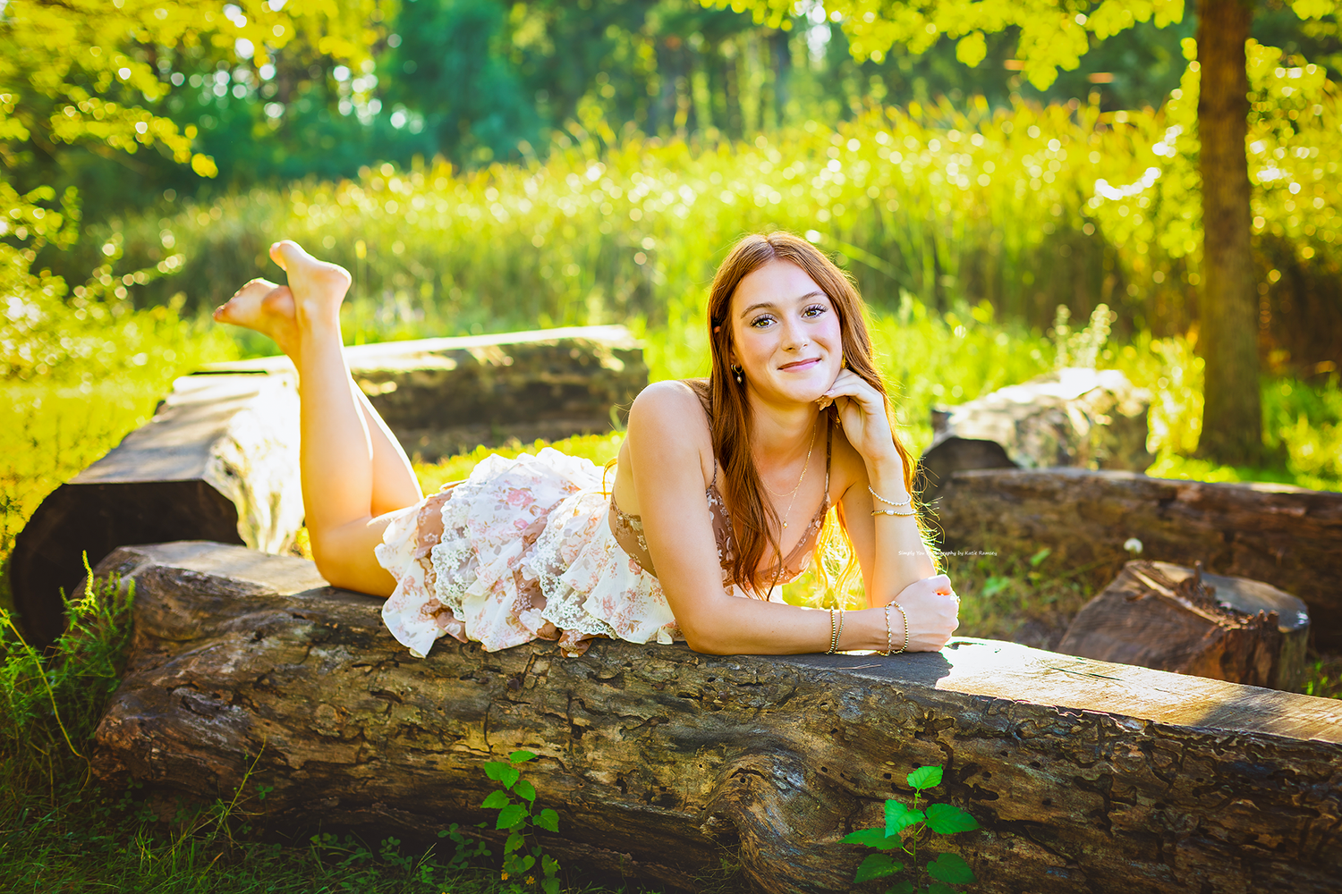 Young woman in floral dress lying on a log in a sunny green park, smiling peacefully at the camera.