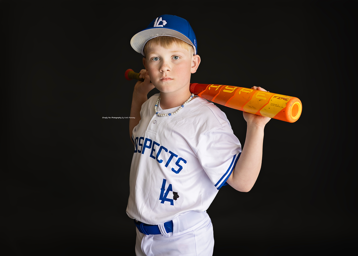 Young boy in Prospects baseball uniform poses with orange bat on shoulder against dark background.