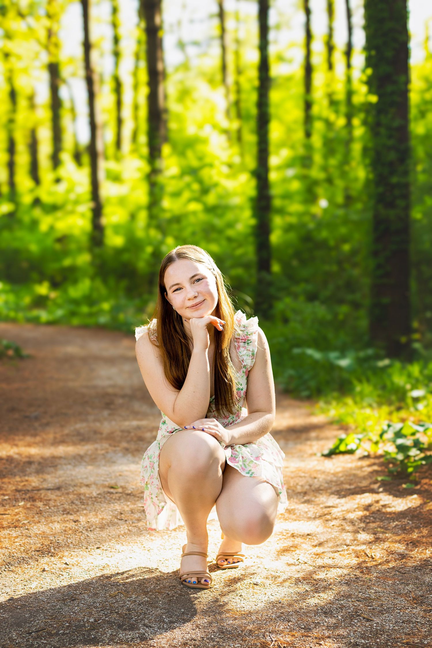 Young woman in floral dress crouching on a sunlit forest path surrounded by lush green trees.
