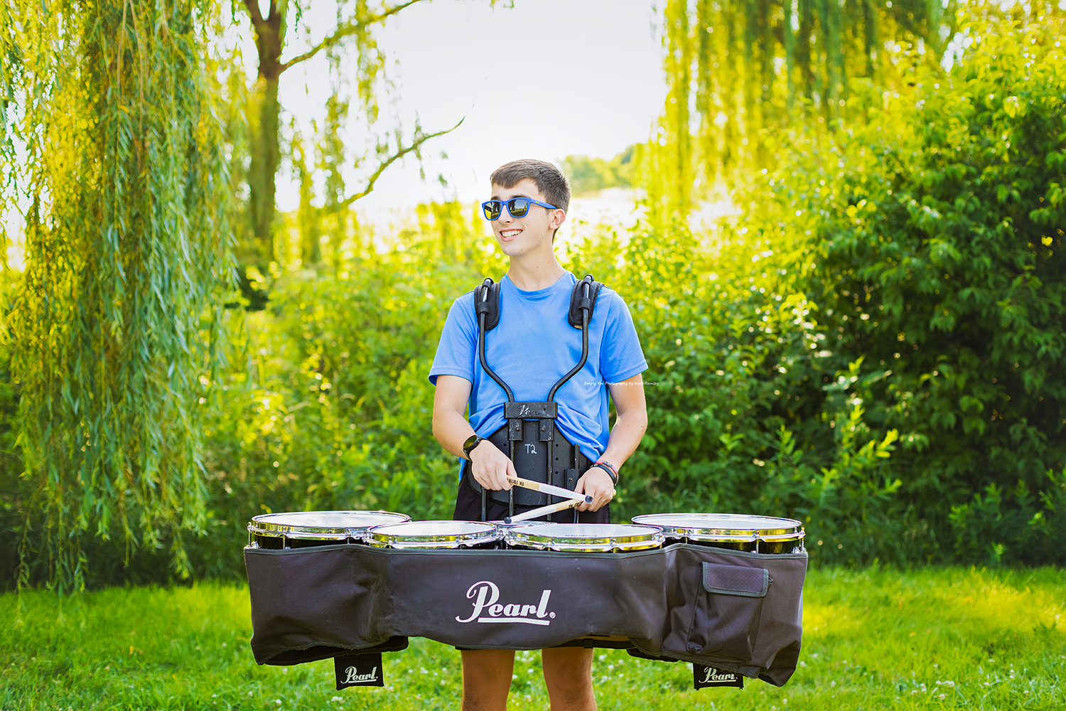 Teen boy in blue shirt and sunglasses playing Pearl tenor drums outdoors in a lush green park setting.