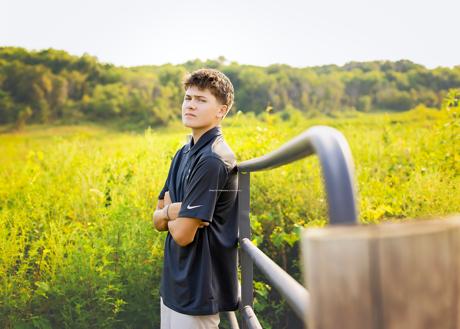 Teen boy in navy Nike shirt leans on metal railing in sunny green meadow for senior portrait photo.