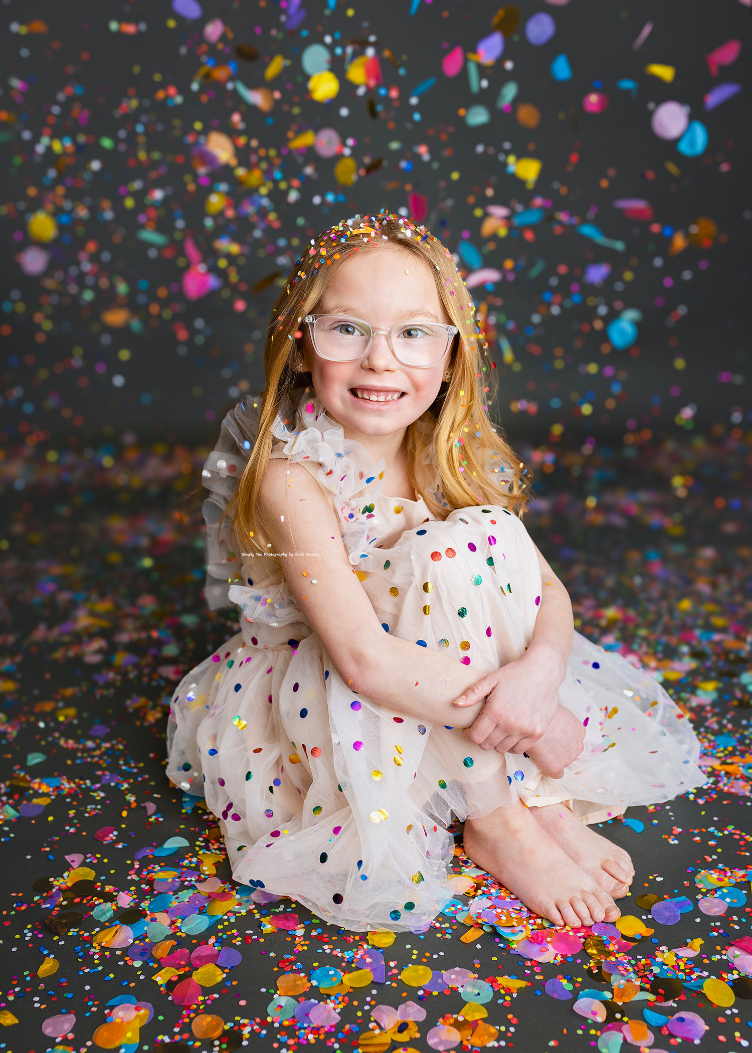 Smiling redhead girl in polka dot dress and glasses sits among colorful confetti falling on dark background.