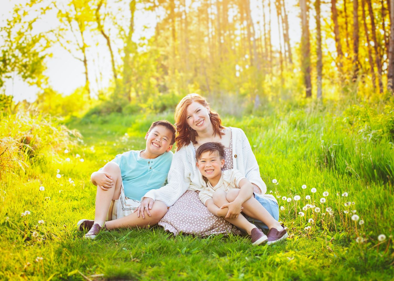 A woman and two boys relax together on green grass in a bright, sunlit outdoor setting.