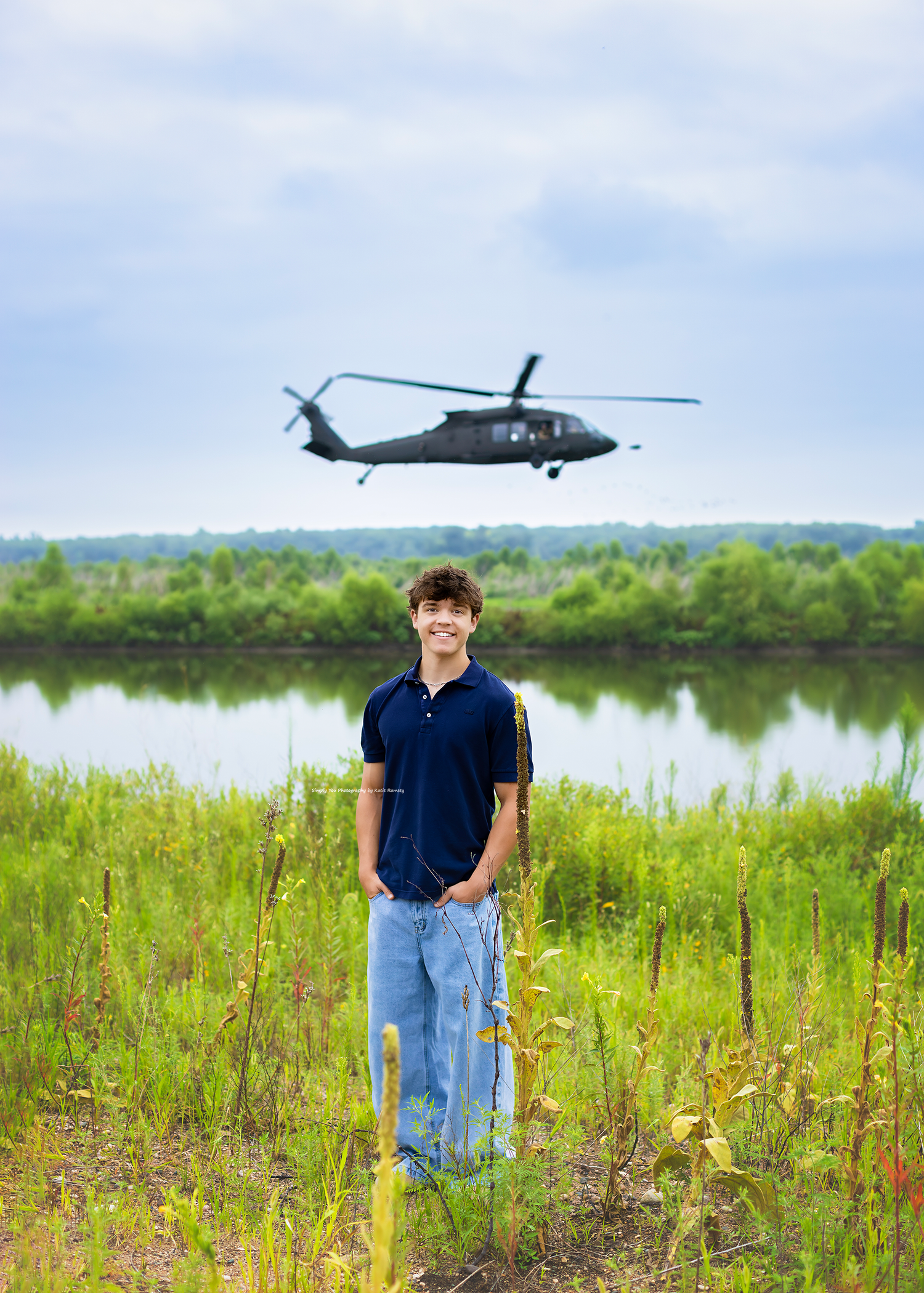 Young man smiling in a field with a military Black Hawk helicopter flying over a river behind him.