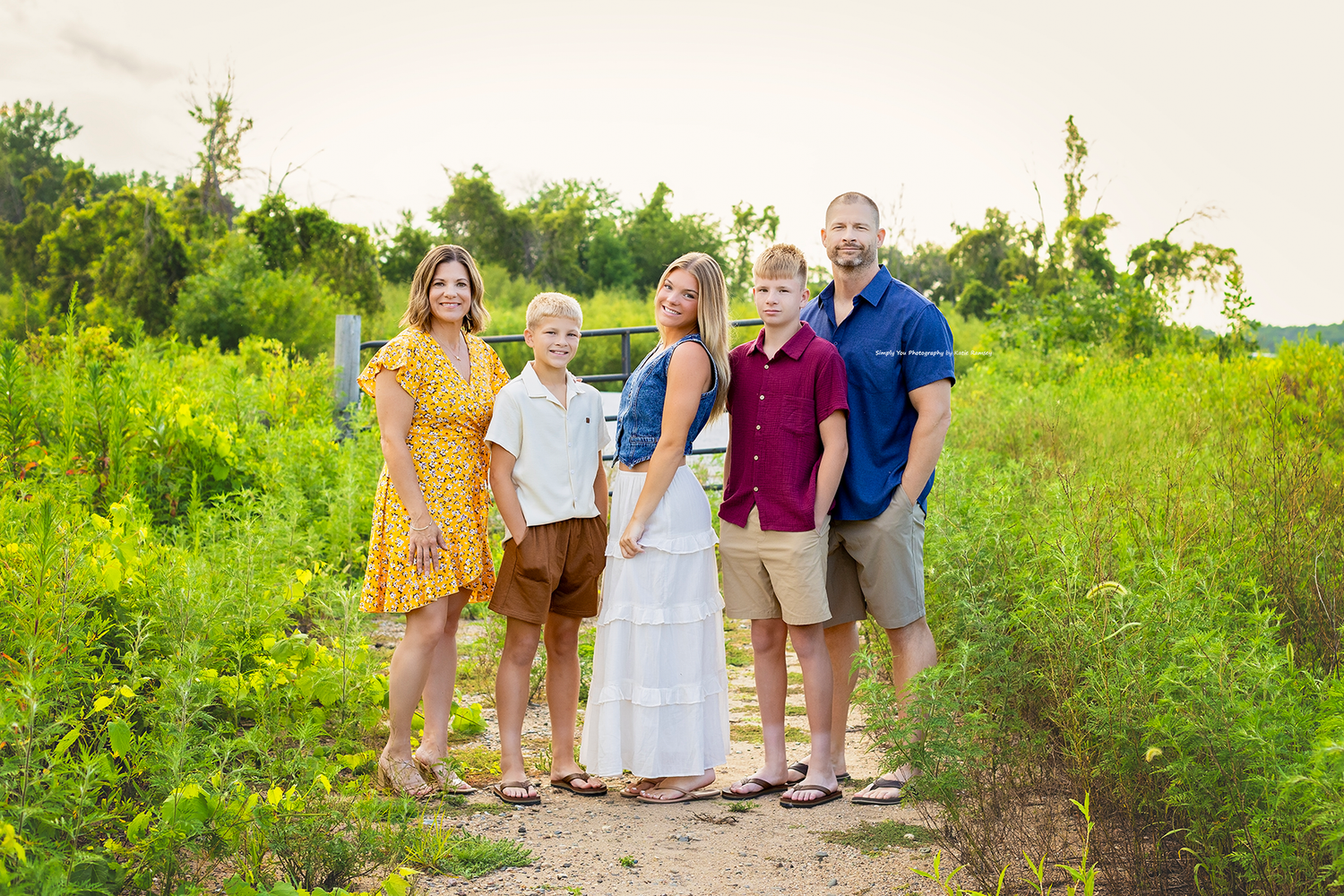 Family of five posing outdoors on a dirt path surrounded by lush green fields during golden hour.