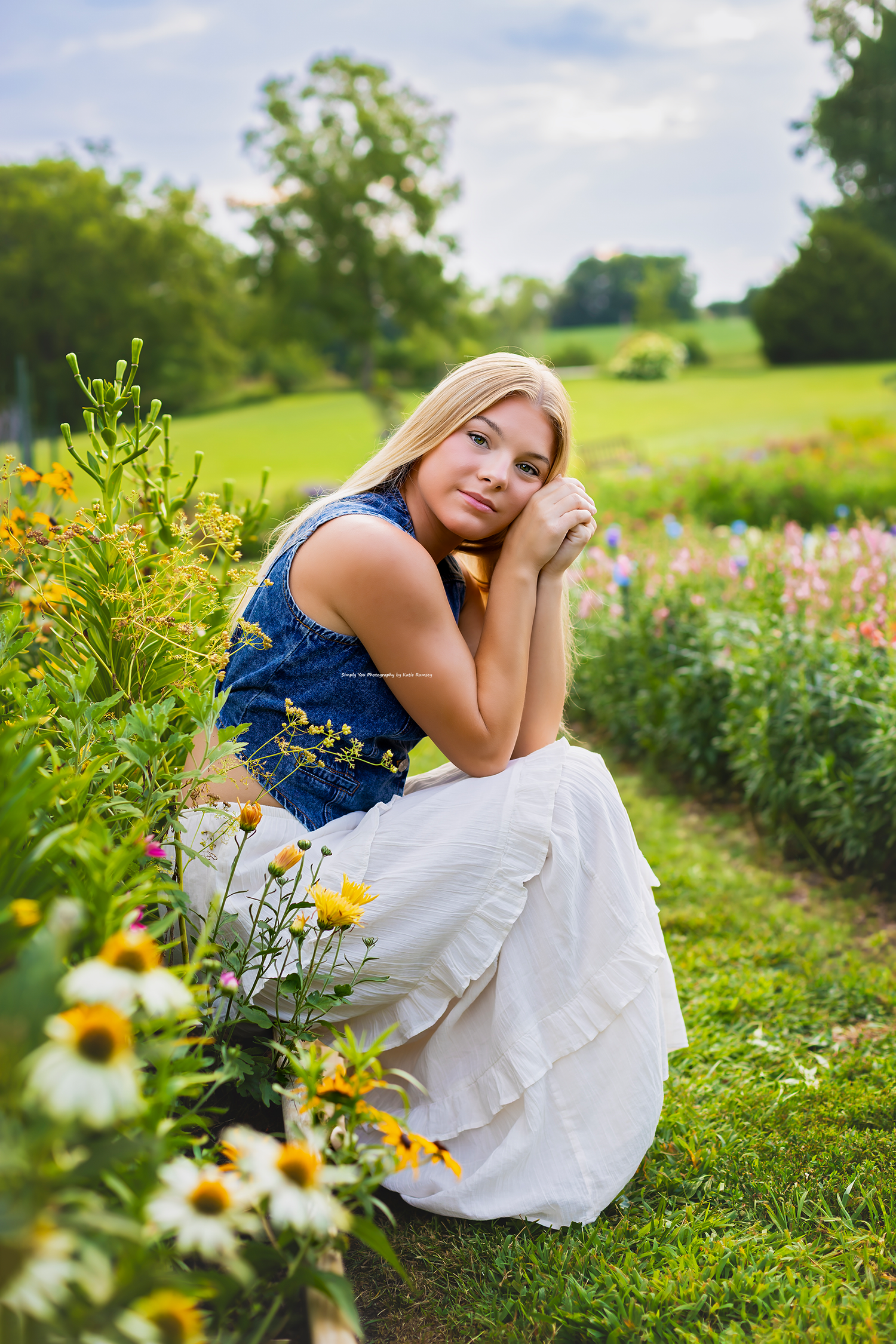 Blonde girl in blue top and white skirt crouching among colorful wildflowers in a lush green garden.