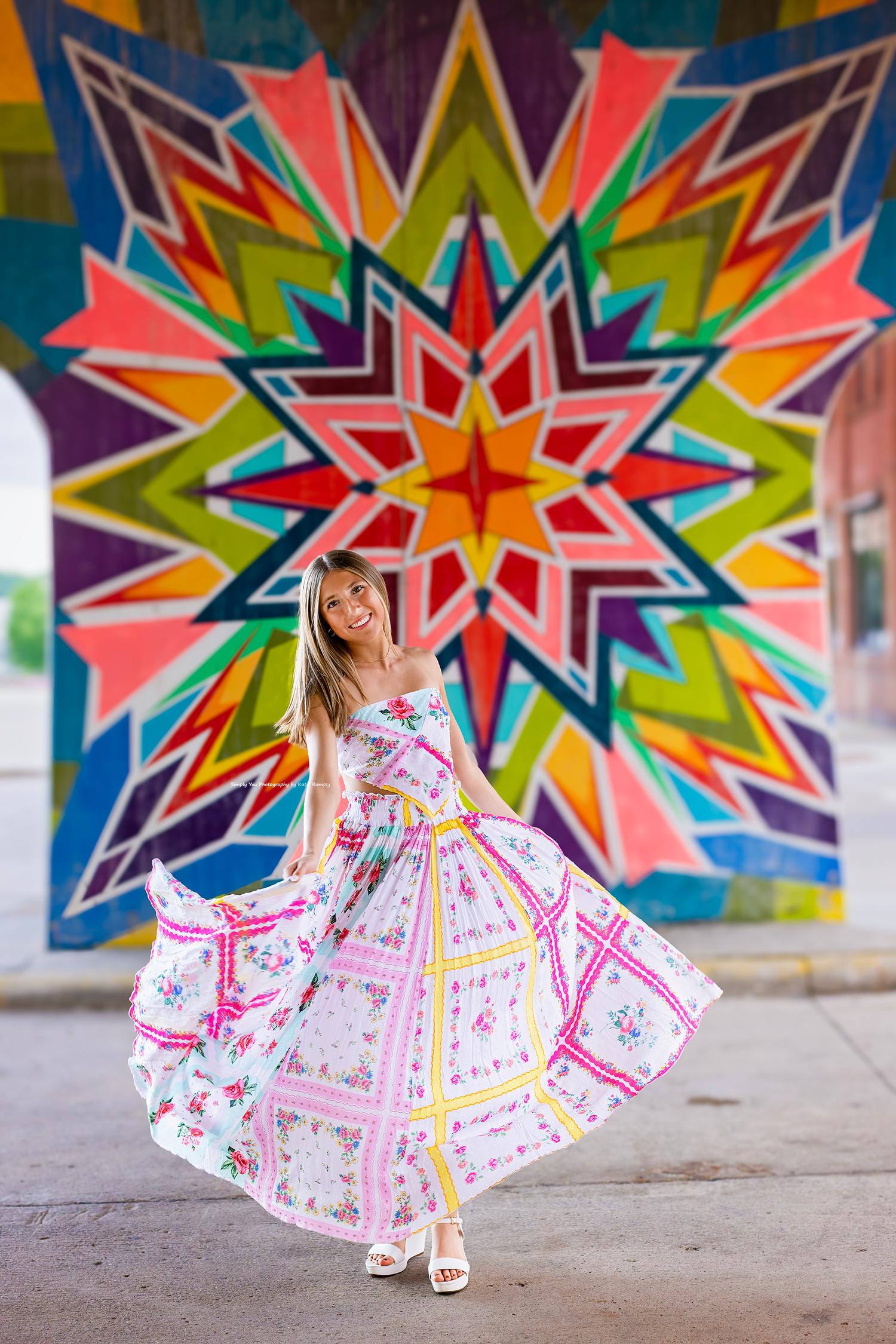 Young woman in floral maxi dress twirling in front of colorful geometric star mural wall.