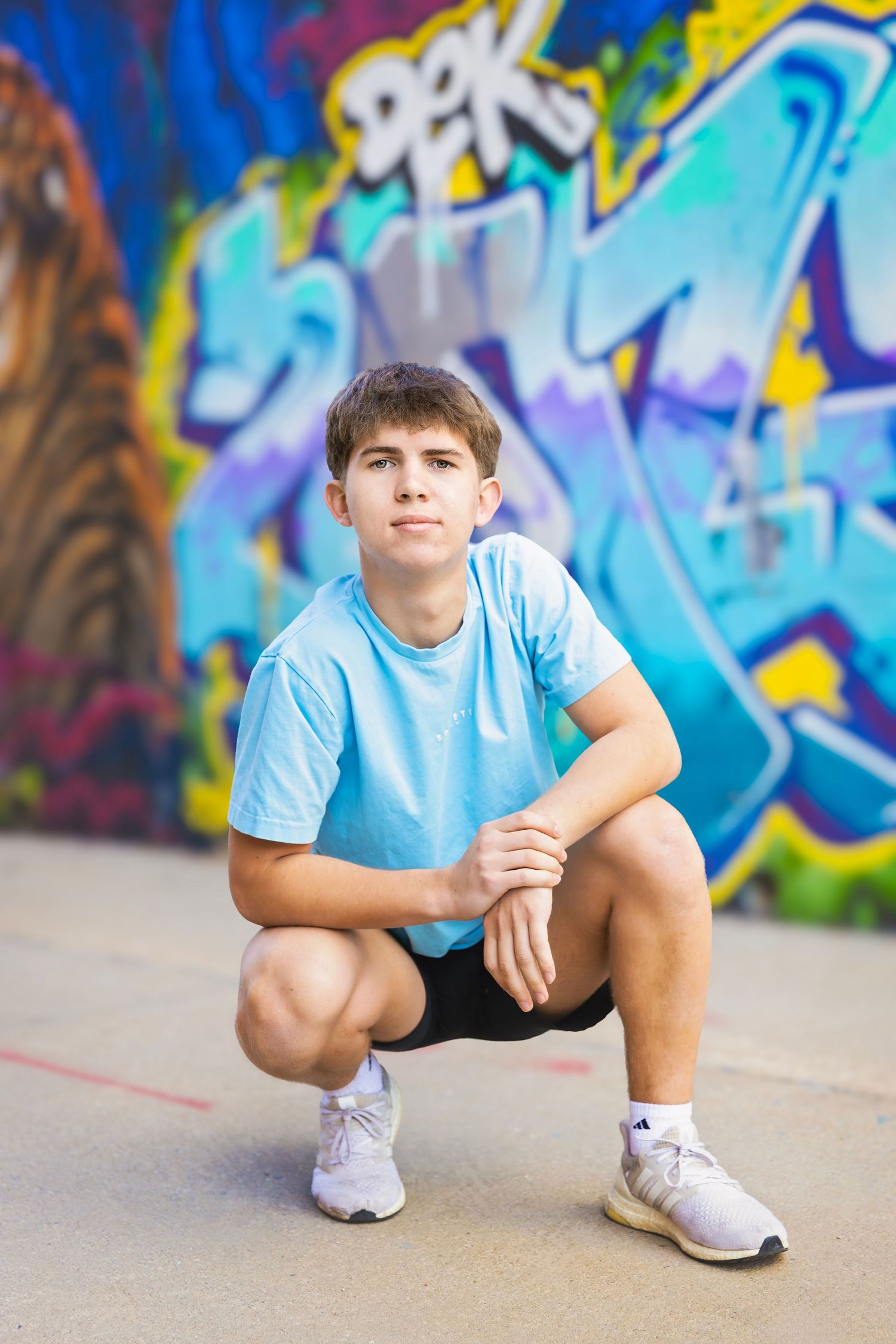 Teen boy in blue shirt crouching in front of colorful graffiti wall outdoors.