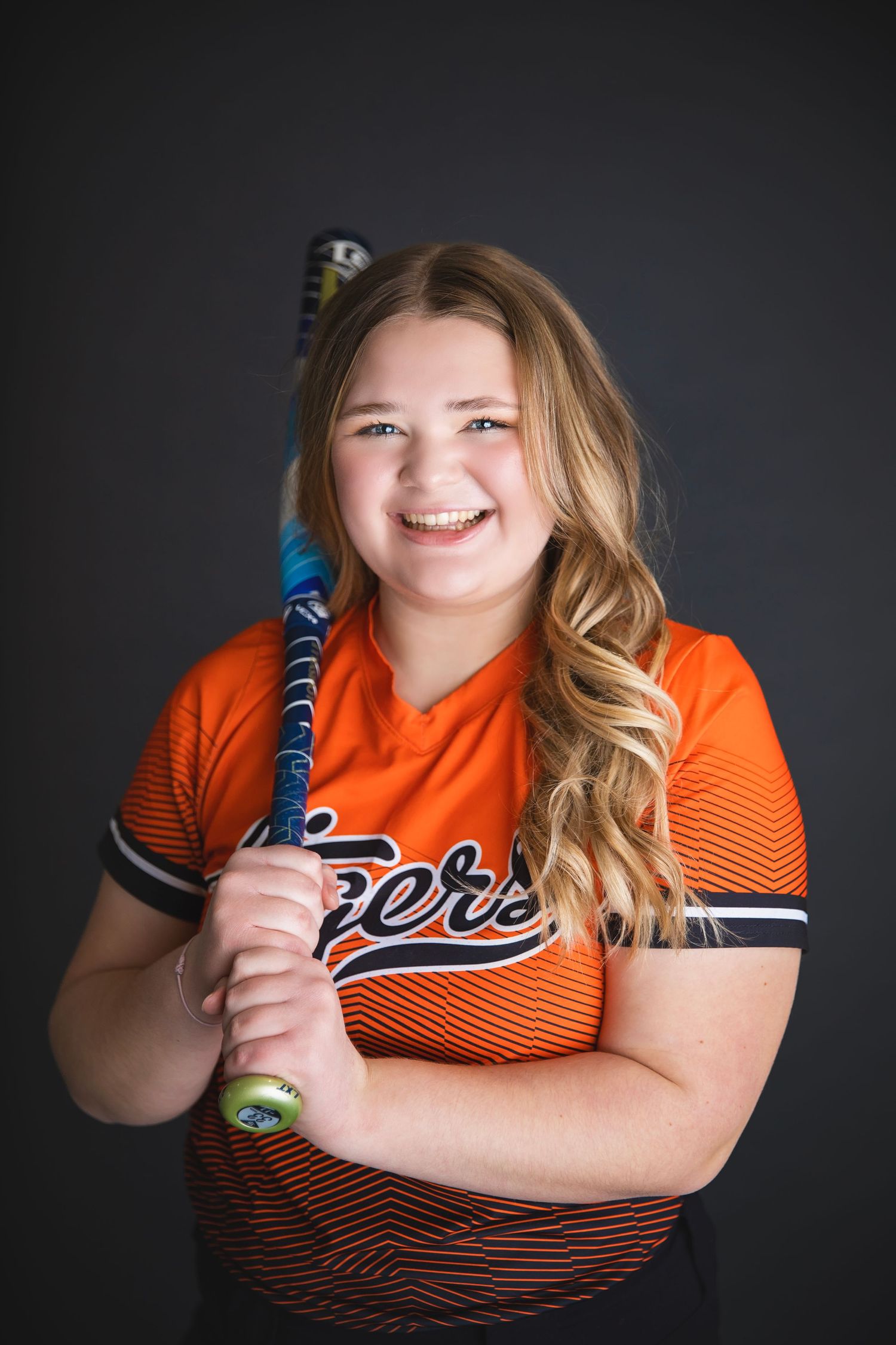 Smiling teen softball player in orange Tigers uniform holds bat in studio portrait photo.