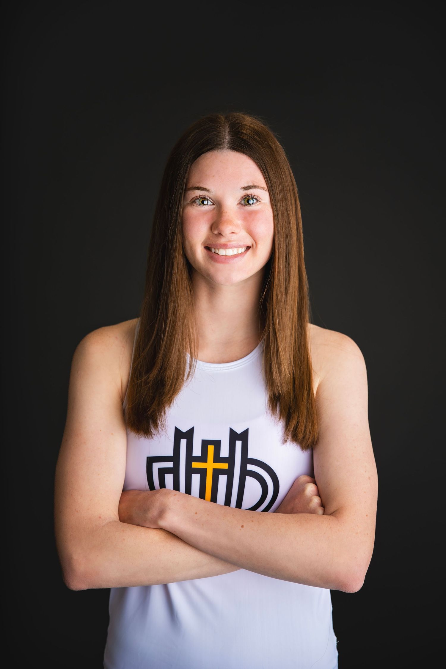Smiling teenage girl in white athletic tank top poses with arms crossed against dark background.