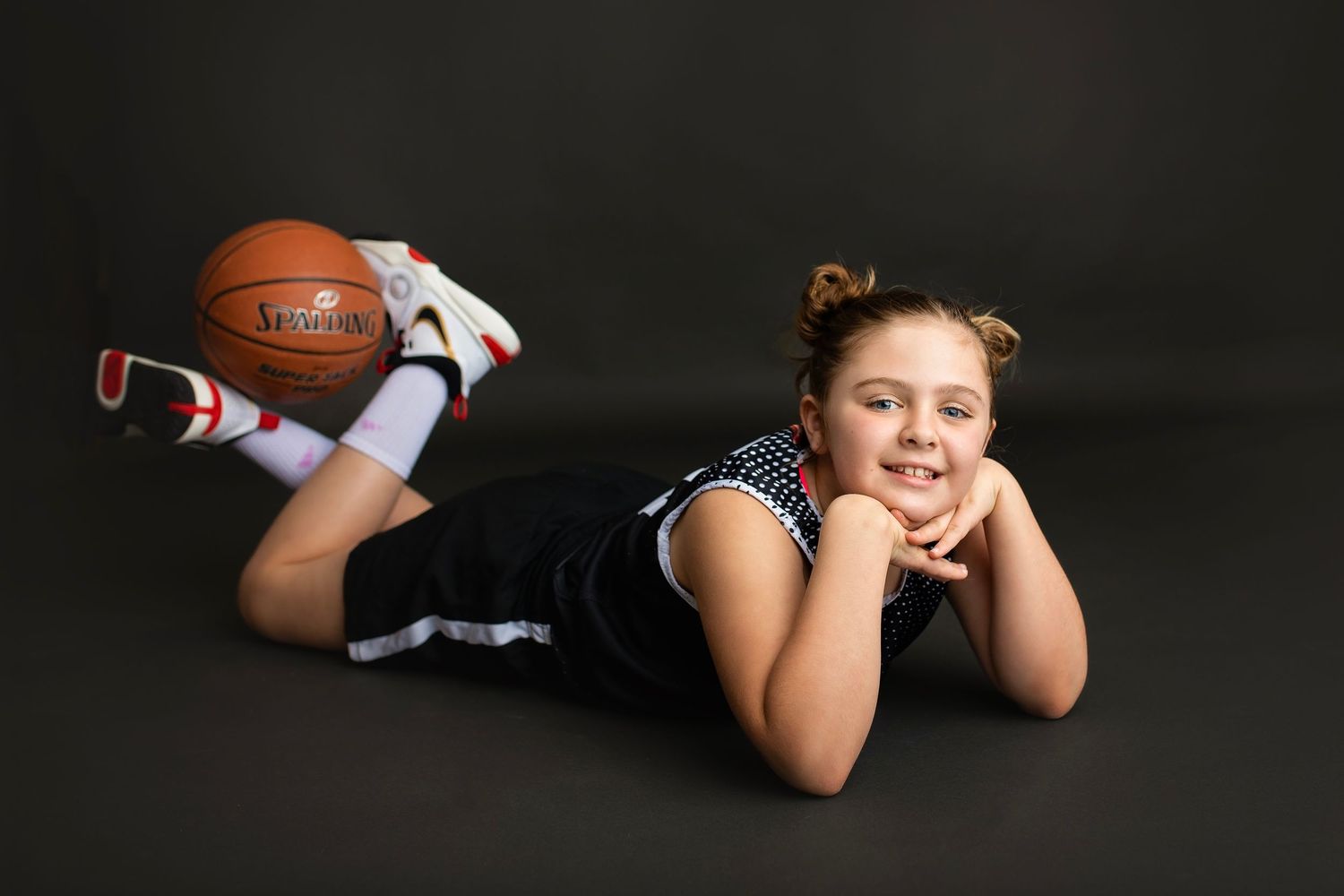 Young girl in black basketball uniform lying on floor smiling with a Spalding basketball behind her.