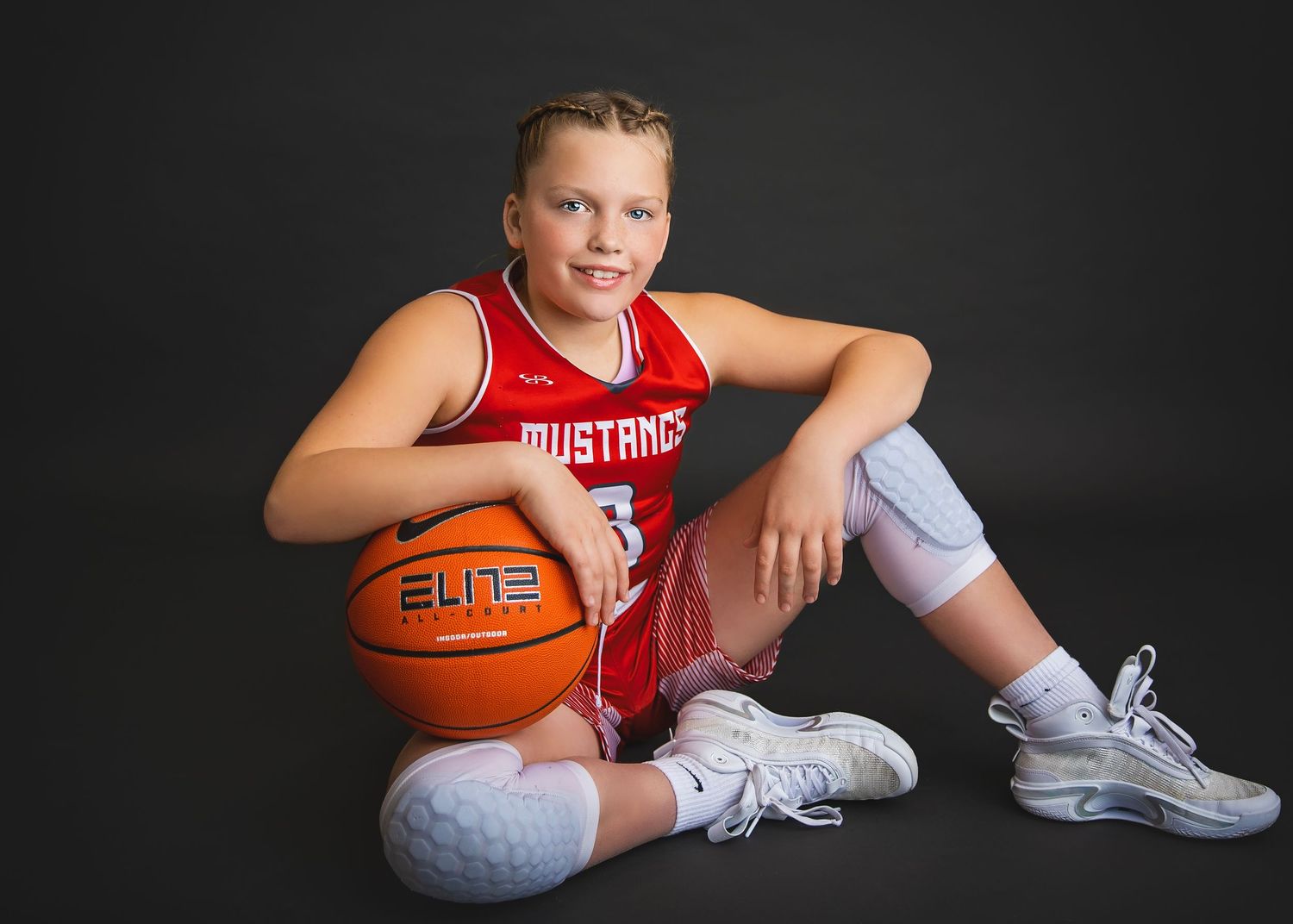 Young girl in red Mustangs basketball jersey sitting with Nike Elite basketball in studio portrait.