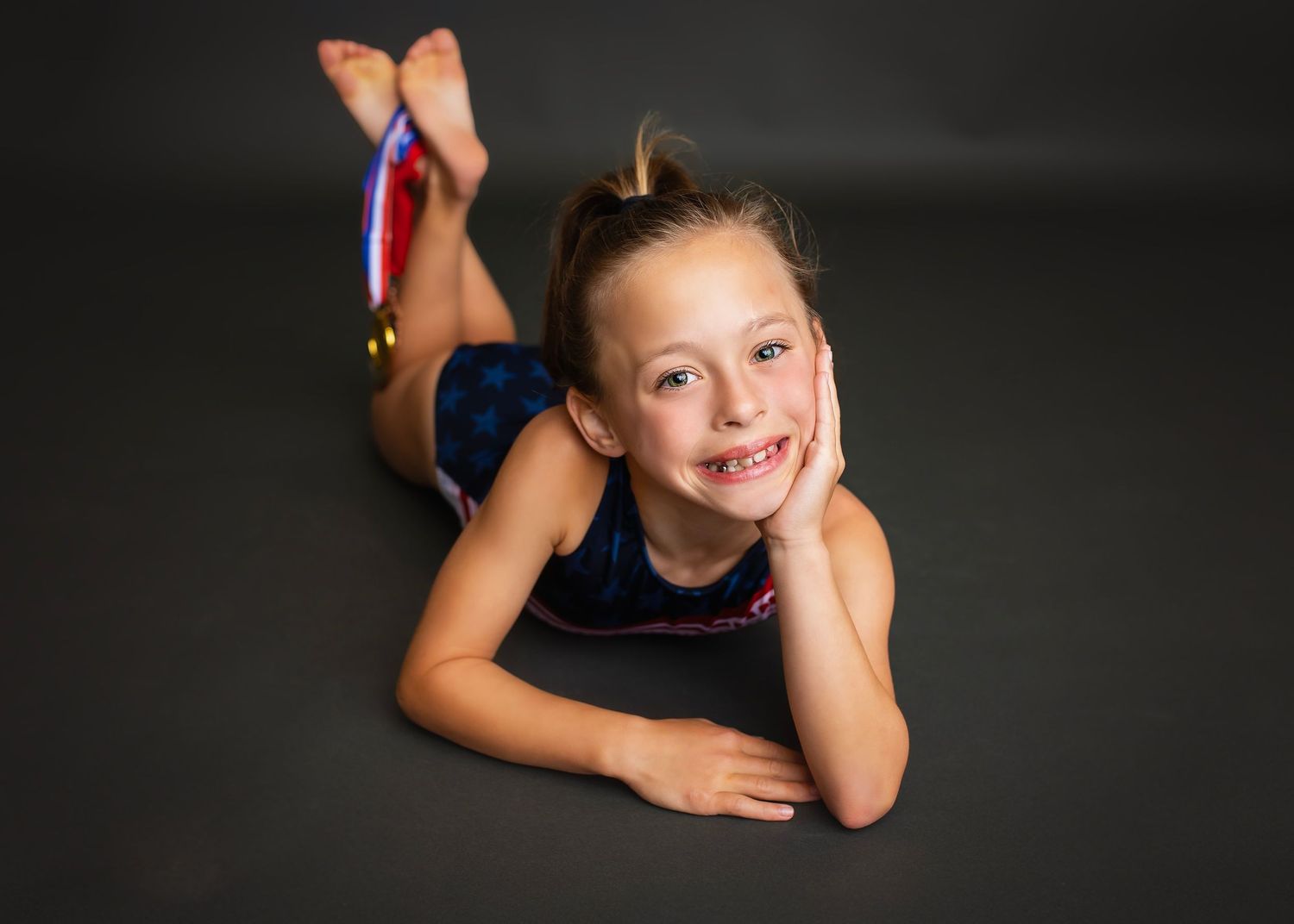 Young smiling gymnast in blue leotard poses lying down wearing a gold medal.